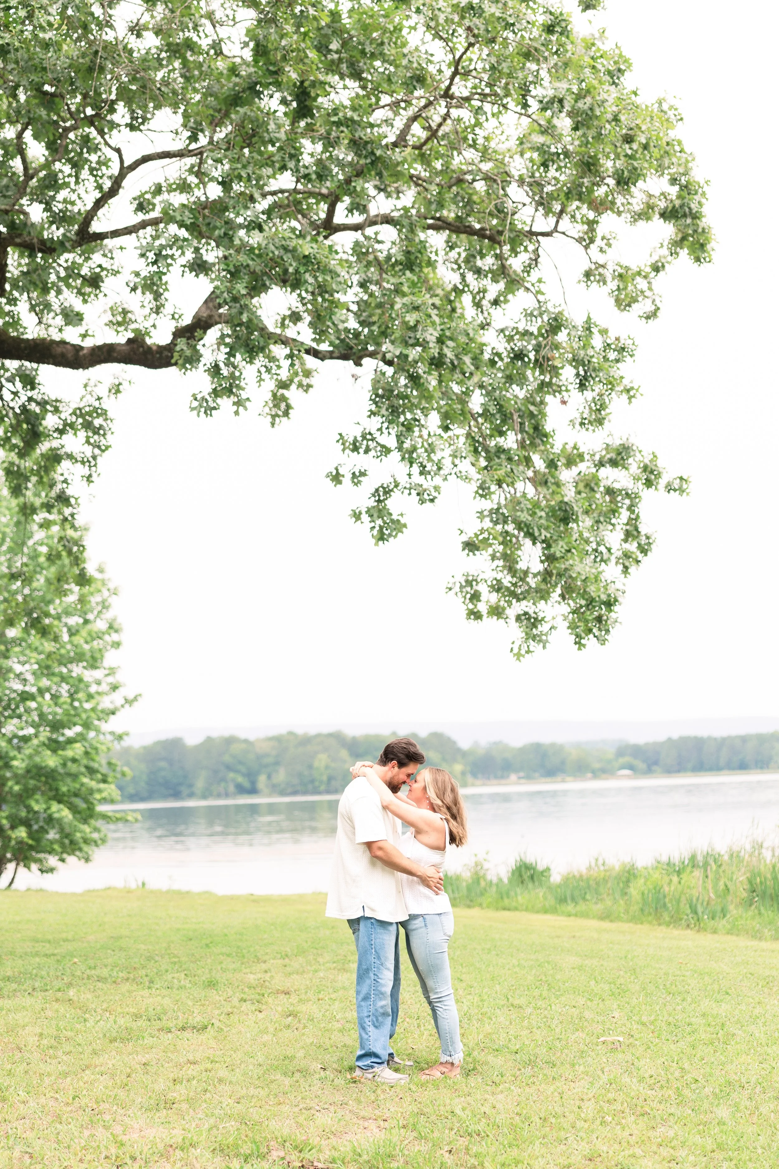 A couple standing close by a lake, hugging under a large tree with broad green leaves, during daytime.