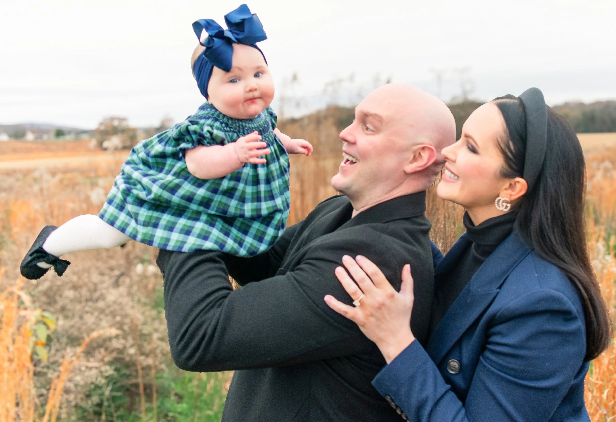 A man holding a baby girl up in the air while a woman looks on smiling in a field during daytime.