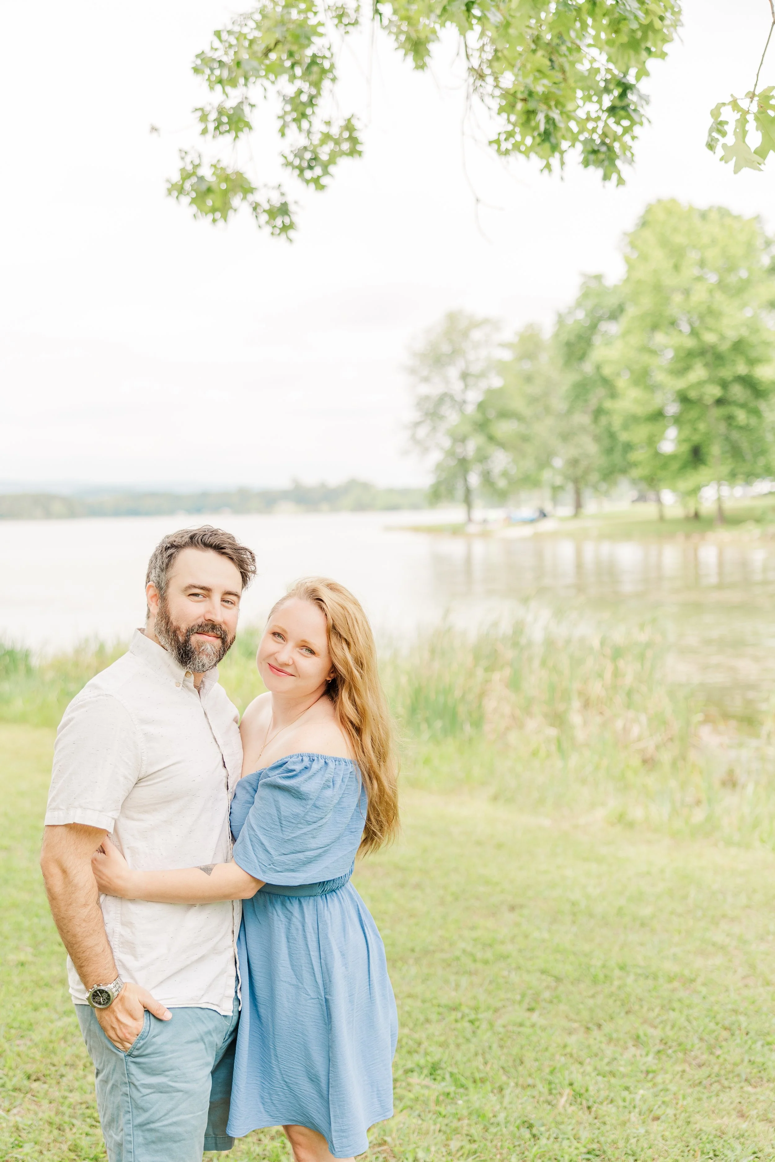 A couple stands closely by a lakeside, smiling with water and trees in the background under a partly cloudy sky.
