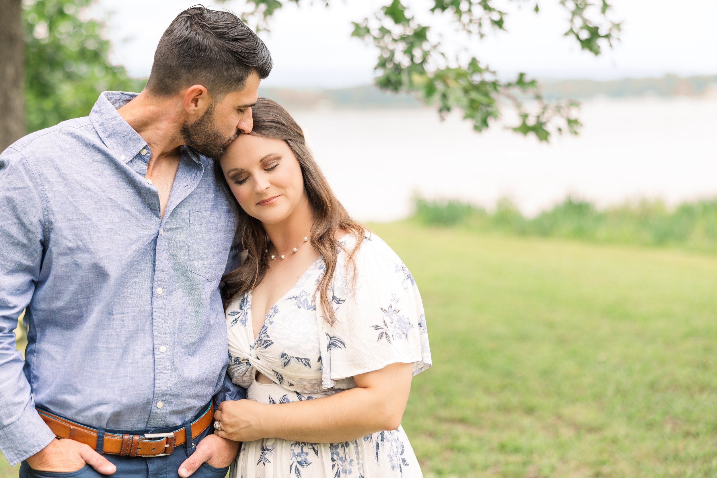 A man and woman standing outdoors near a lake with trees in the background, the man kissing the top of the woman's head with his eyes closed, both with peaceful expressions.