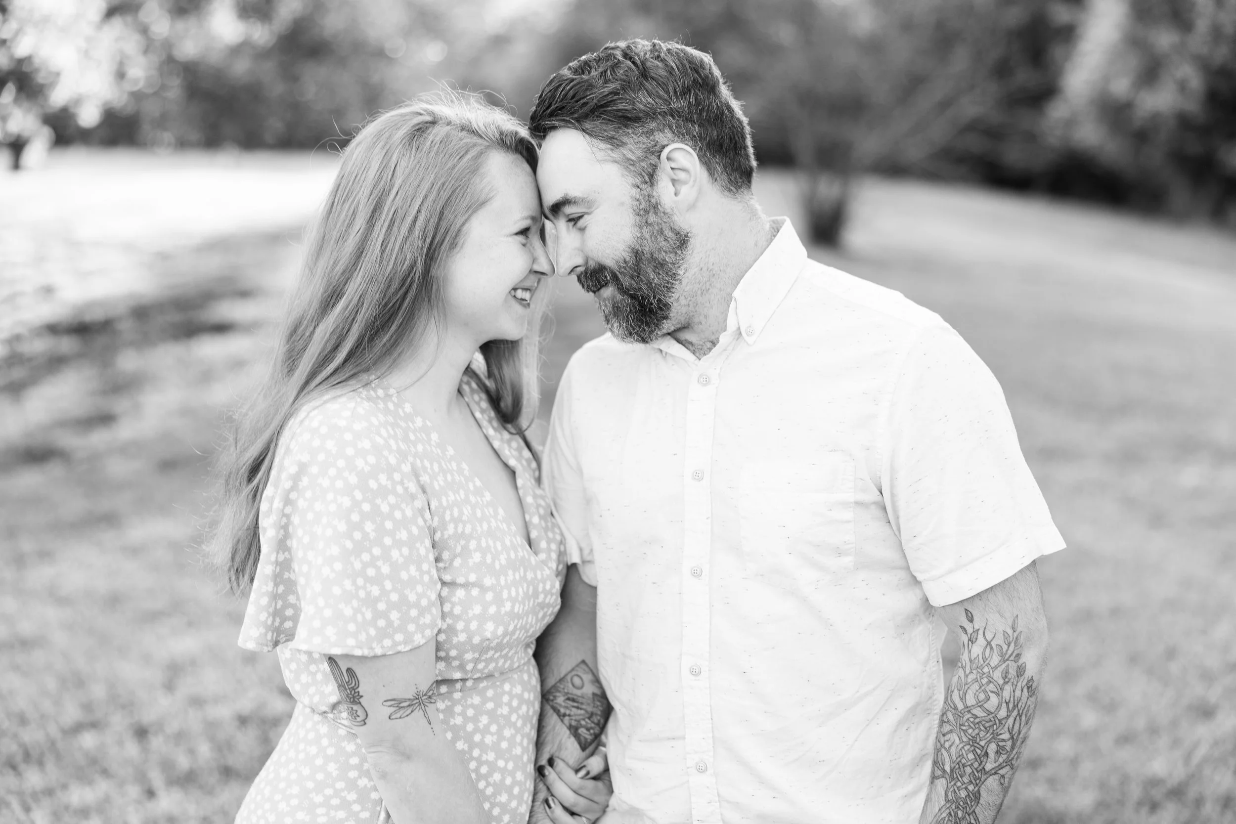 A black and white photo of a couple standing outdoors with their foreheads touching, smiling and looking into each other's eyes, holding hands, with trees and grass in the background.
