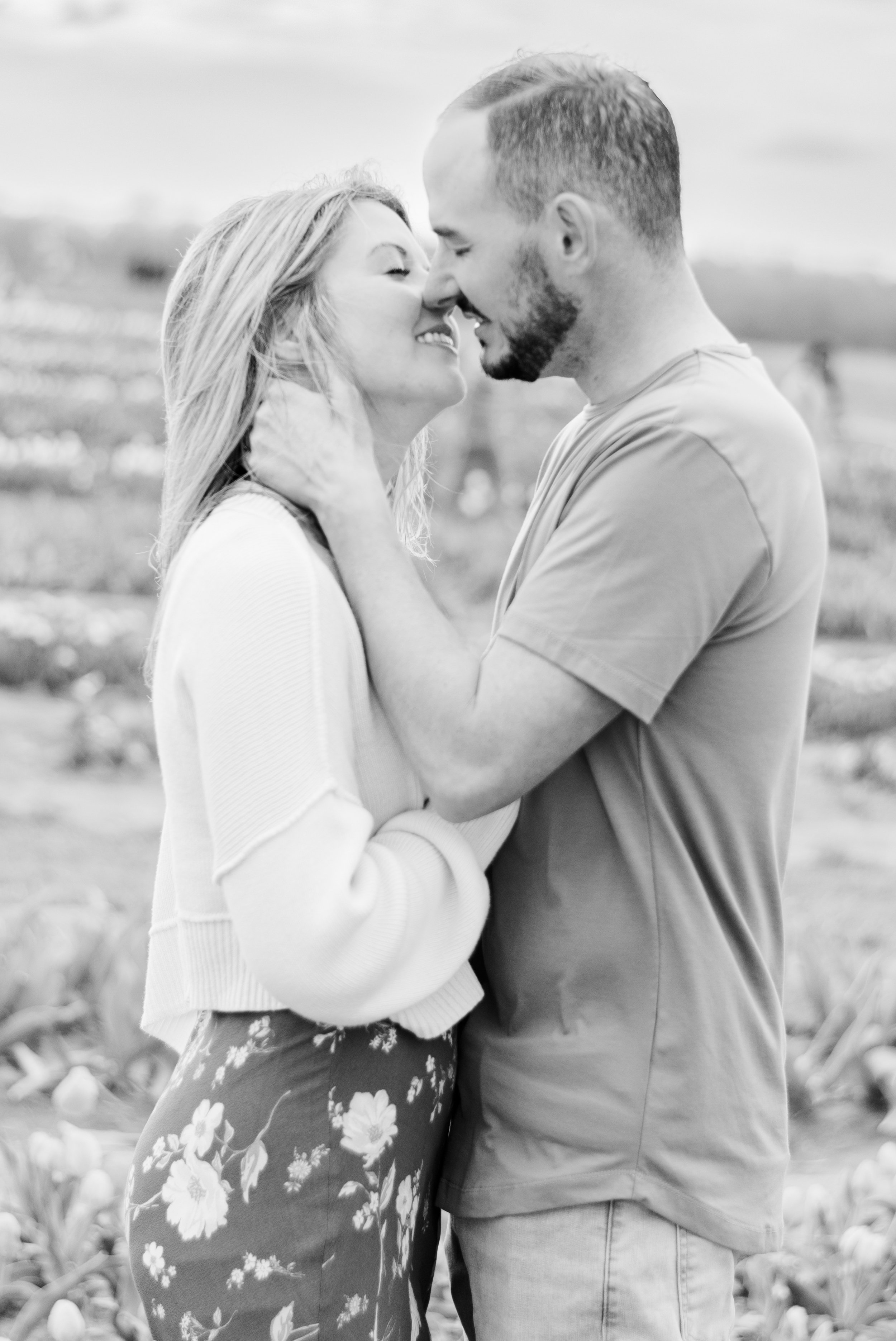A black and white photo of a couple close together, smiling and touching noses in an outdoor setting with flowers in the background.