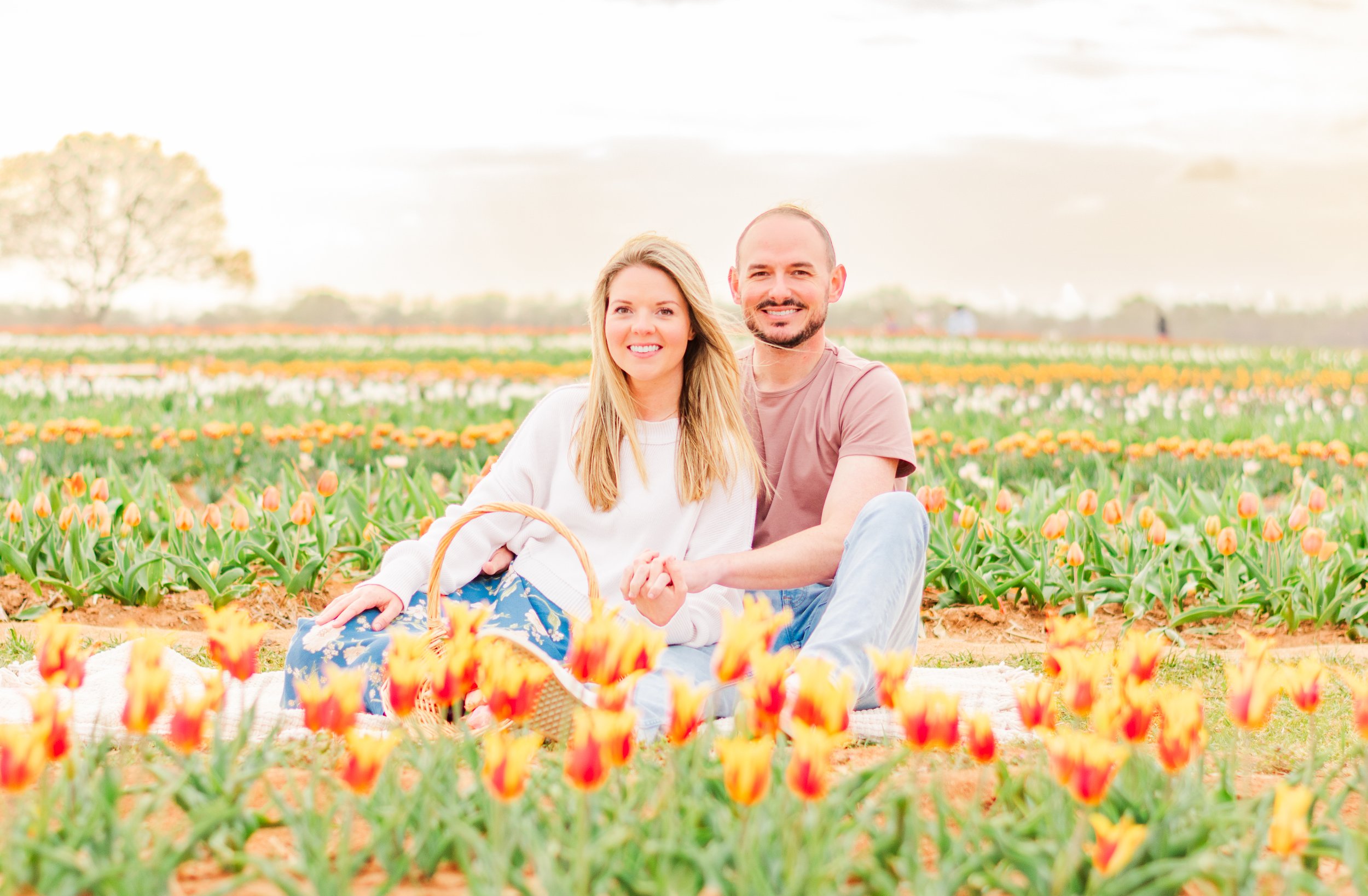 A smiling couple sitting on a blanket in a tulip field with colorful tulips and a cloudy sky.