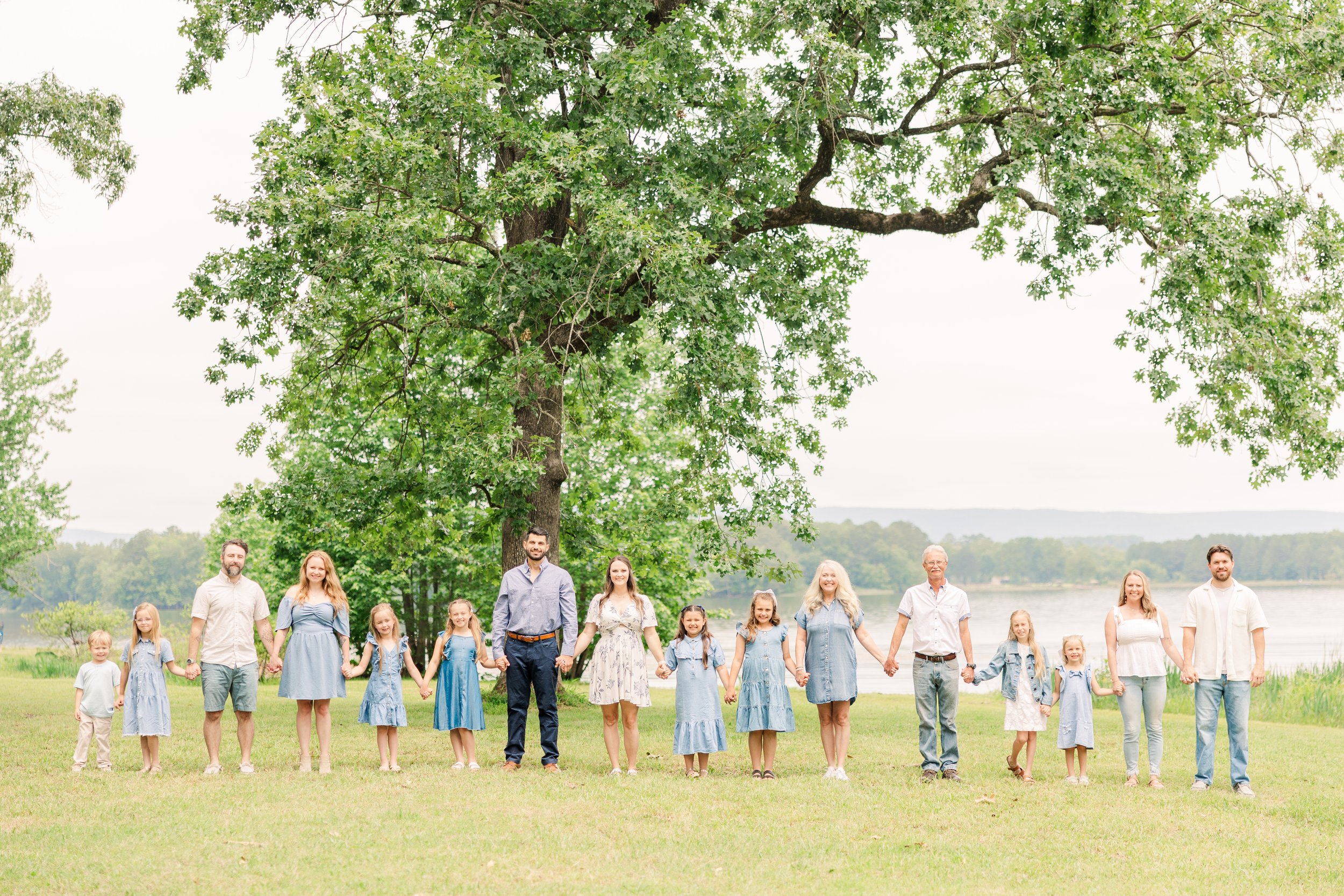 Family standing in a line holding hands outdoors near a large tree and a body of water, smiling on a bright day.