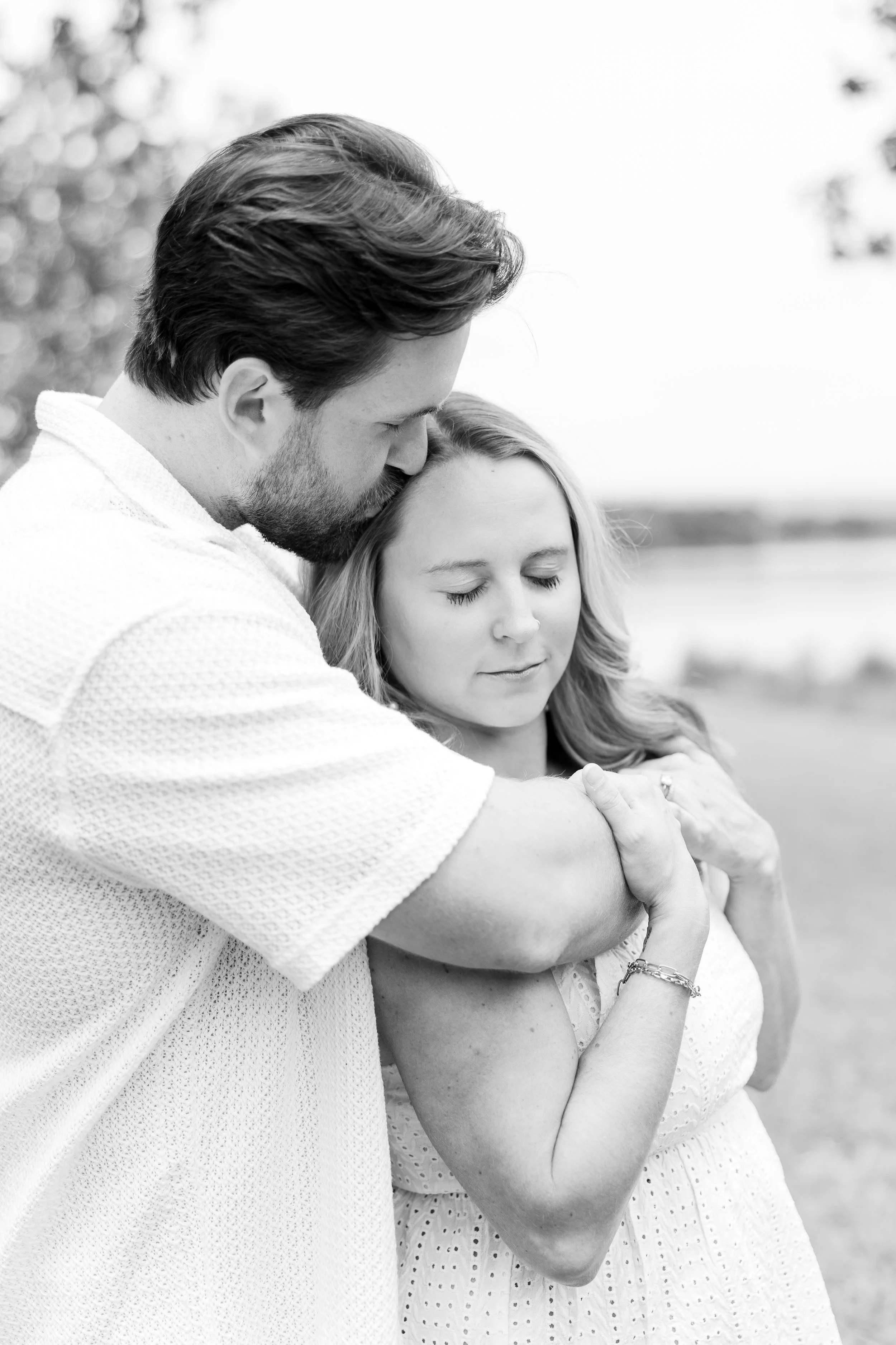 A black and white photo of a man hugging a woman with his head resting on her forehead, outdoors near a body of water.
