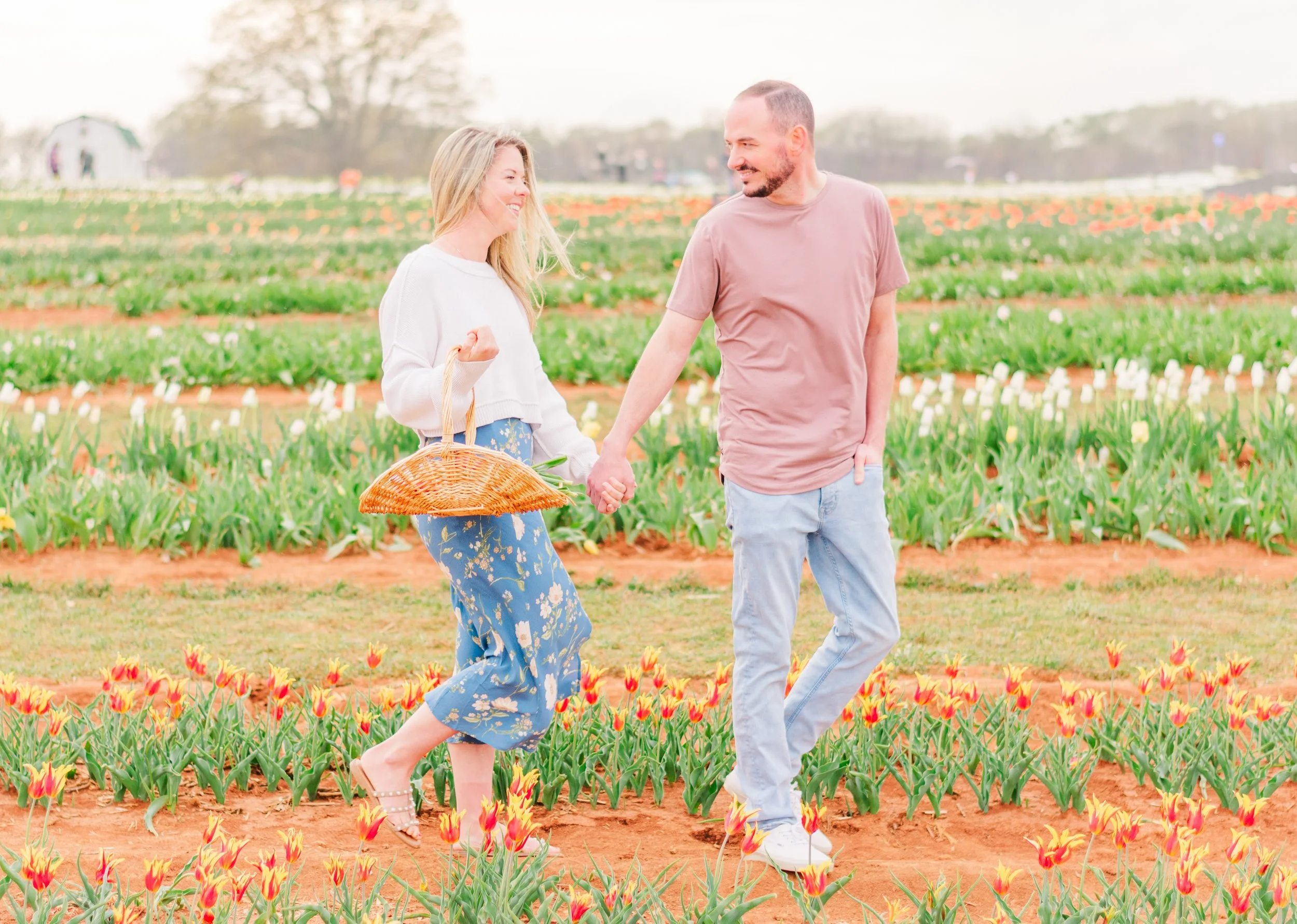 A couple holding hands and smiling in a tulip field during daytime, with the woman carrying a large straw basket.