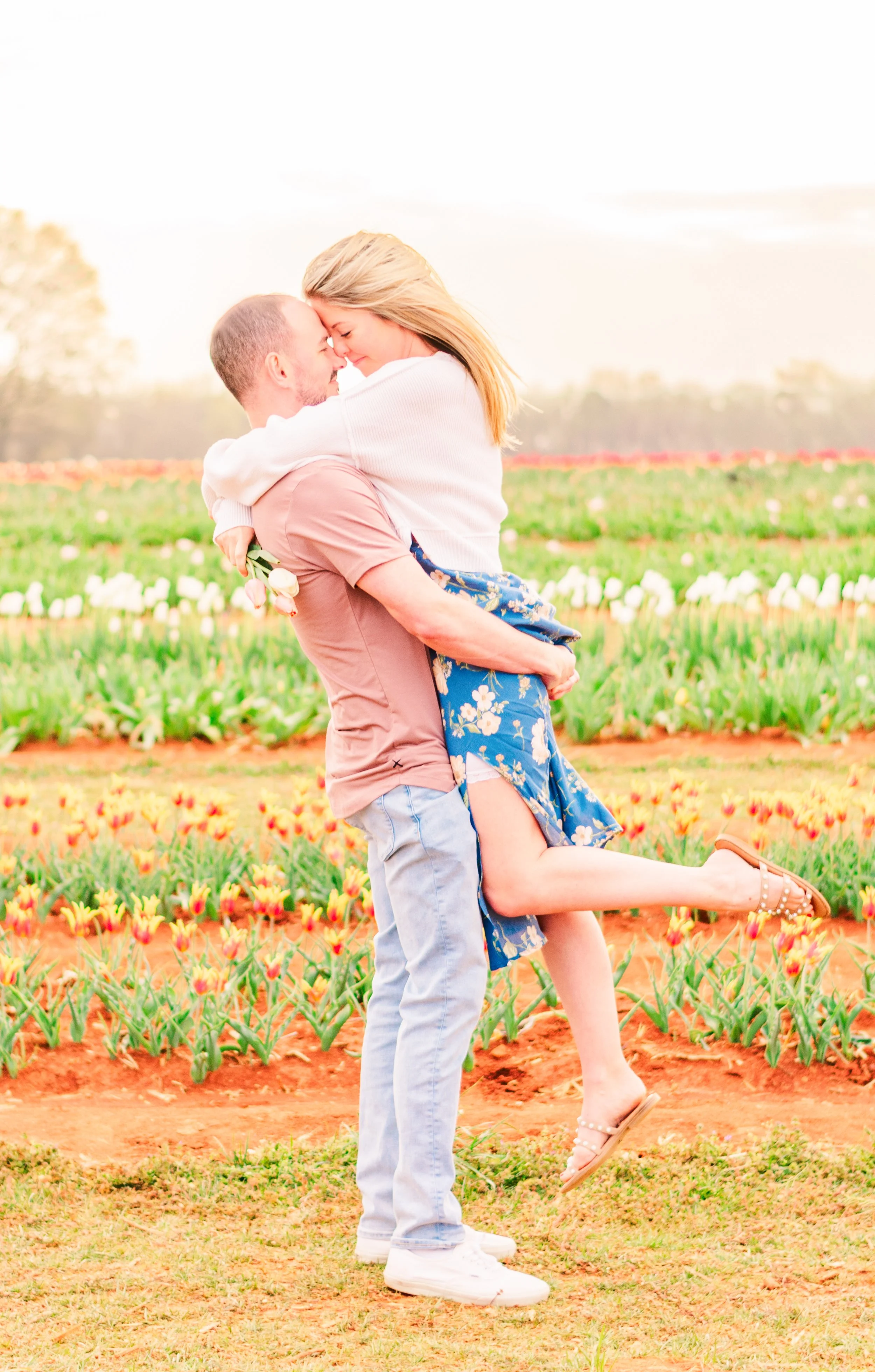 A couple hugging in a tulip field during daytime, the man lifting the woman off the ground as they hold each other close, both smiling with noses touching.