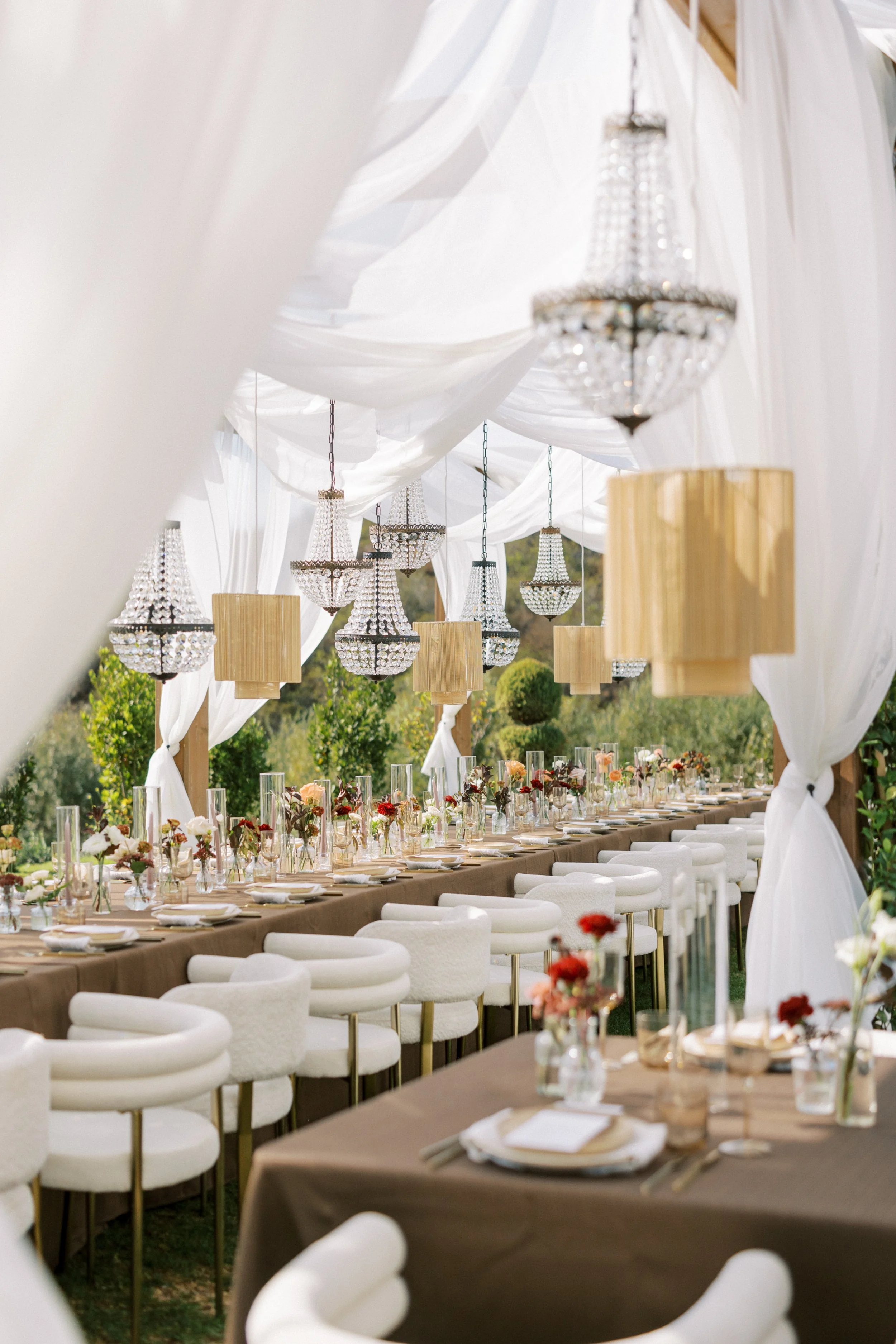 Elegant outdoor dining setup with long table, white chairs, floral centerpieces, chandeliers, and draped white fabric.