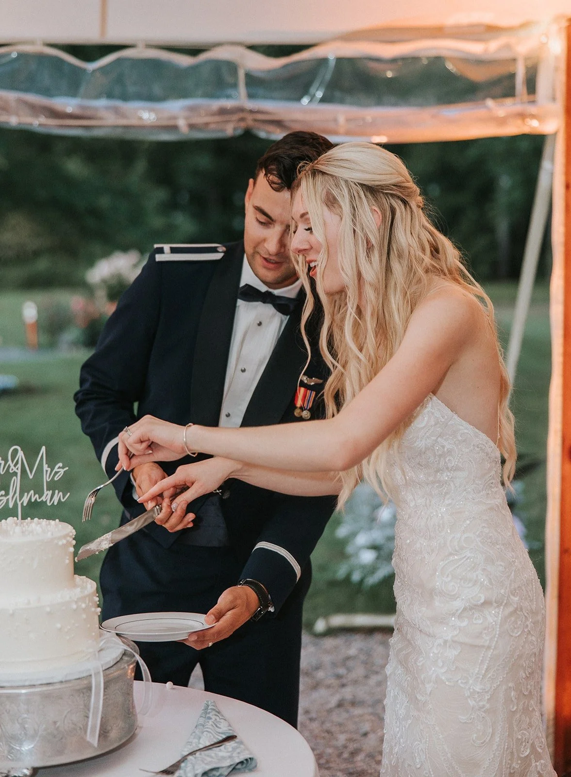 A bride in a white lace wedding dress and a groom in a formal black tuxedo are cutting a wedding cake together, smiling and celebrating outdoors.