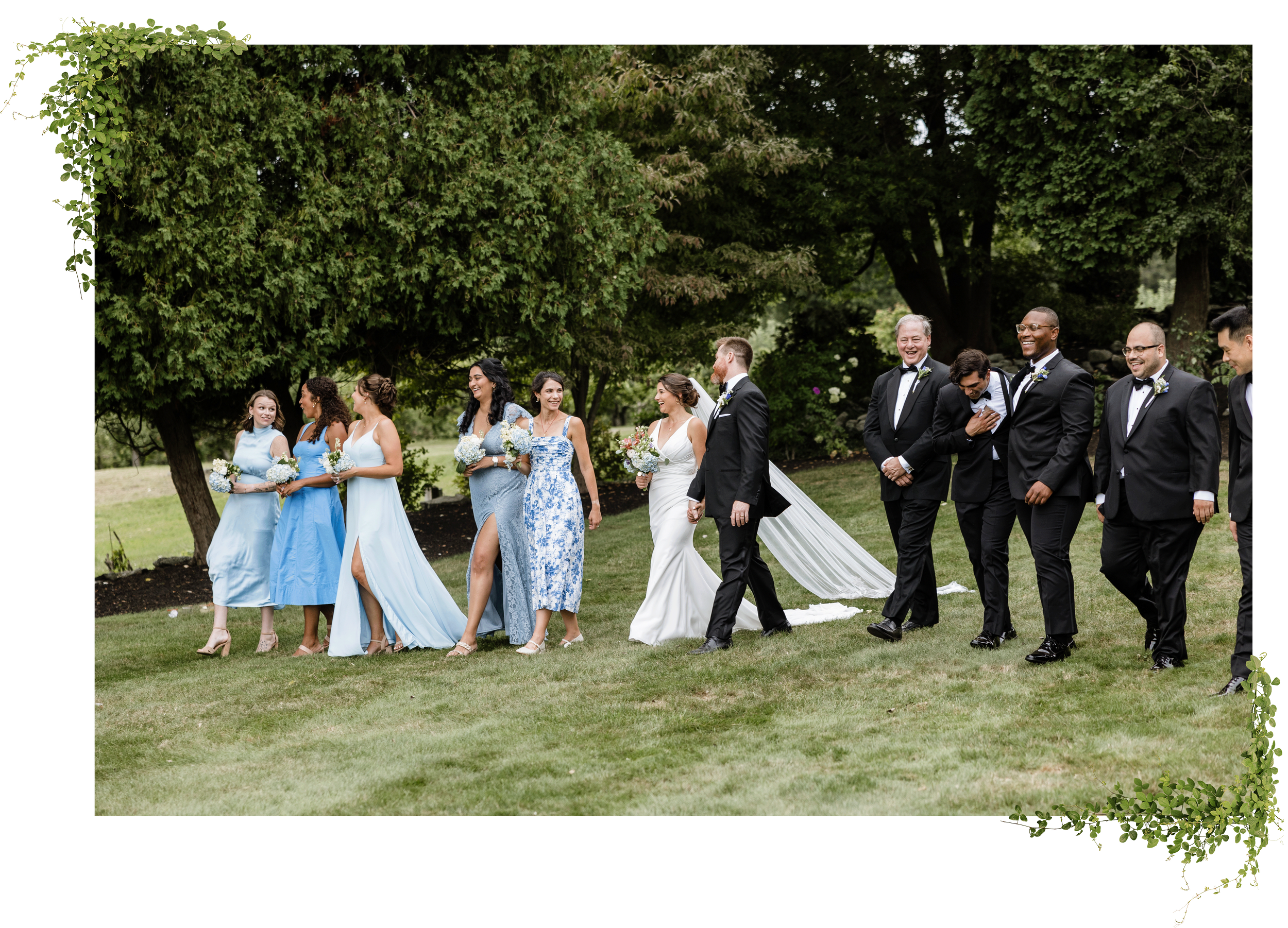 A wedding party walking outdoors on grass with trees in the background. The group consists of bridesmaids in blue dresses, a bride in a white gown, and groomsmen in tuxedos. They are smiling and enjoying the moment.