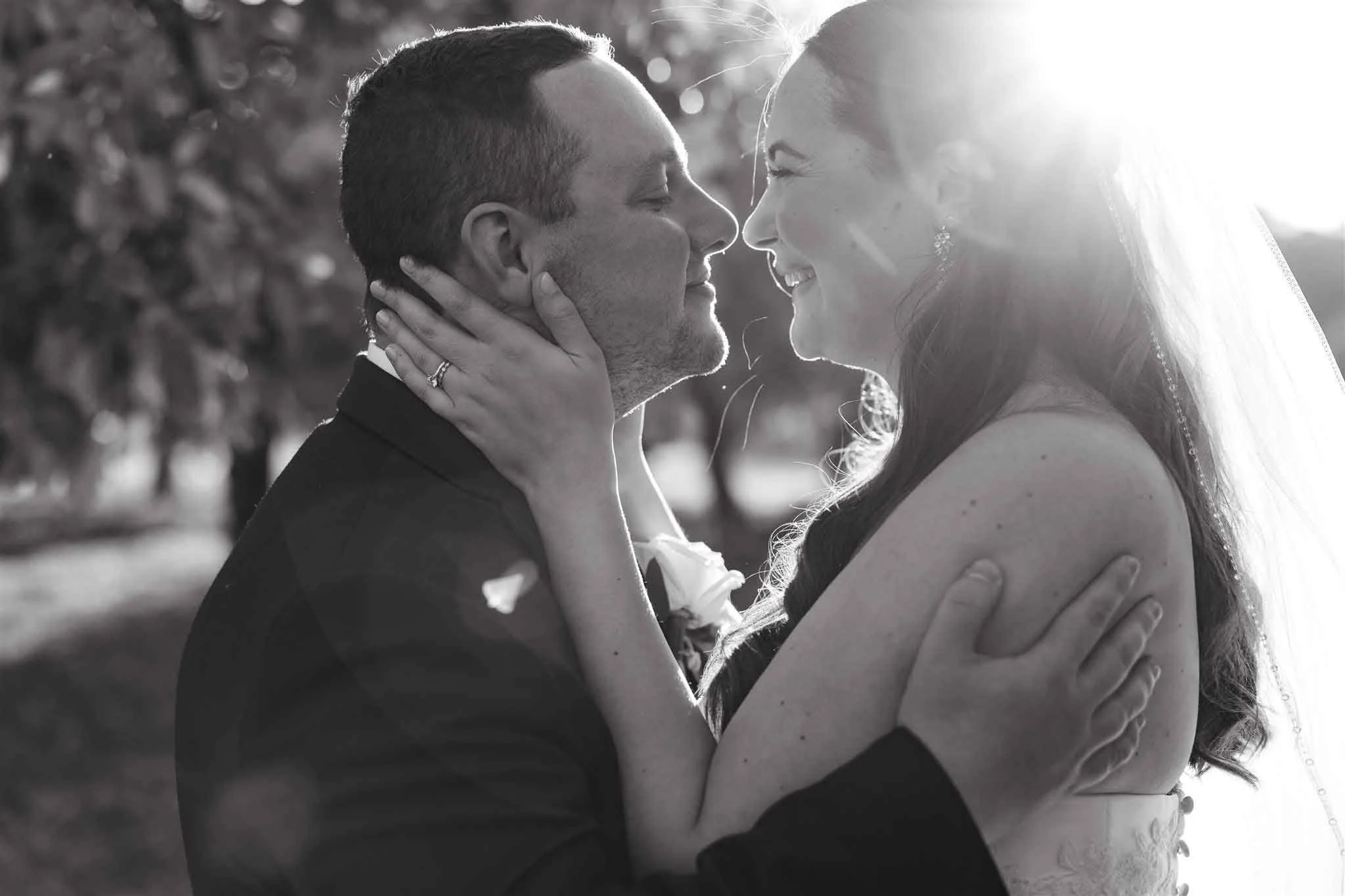 A black and white photo of a wedding couple outdoors, facing each other closely, with the man touching the woman's face and the woman holding the man's neck, both smiling with their eyes closed as sunlight shines behind them.
