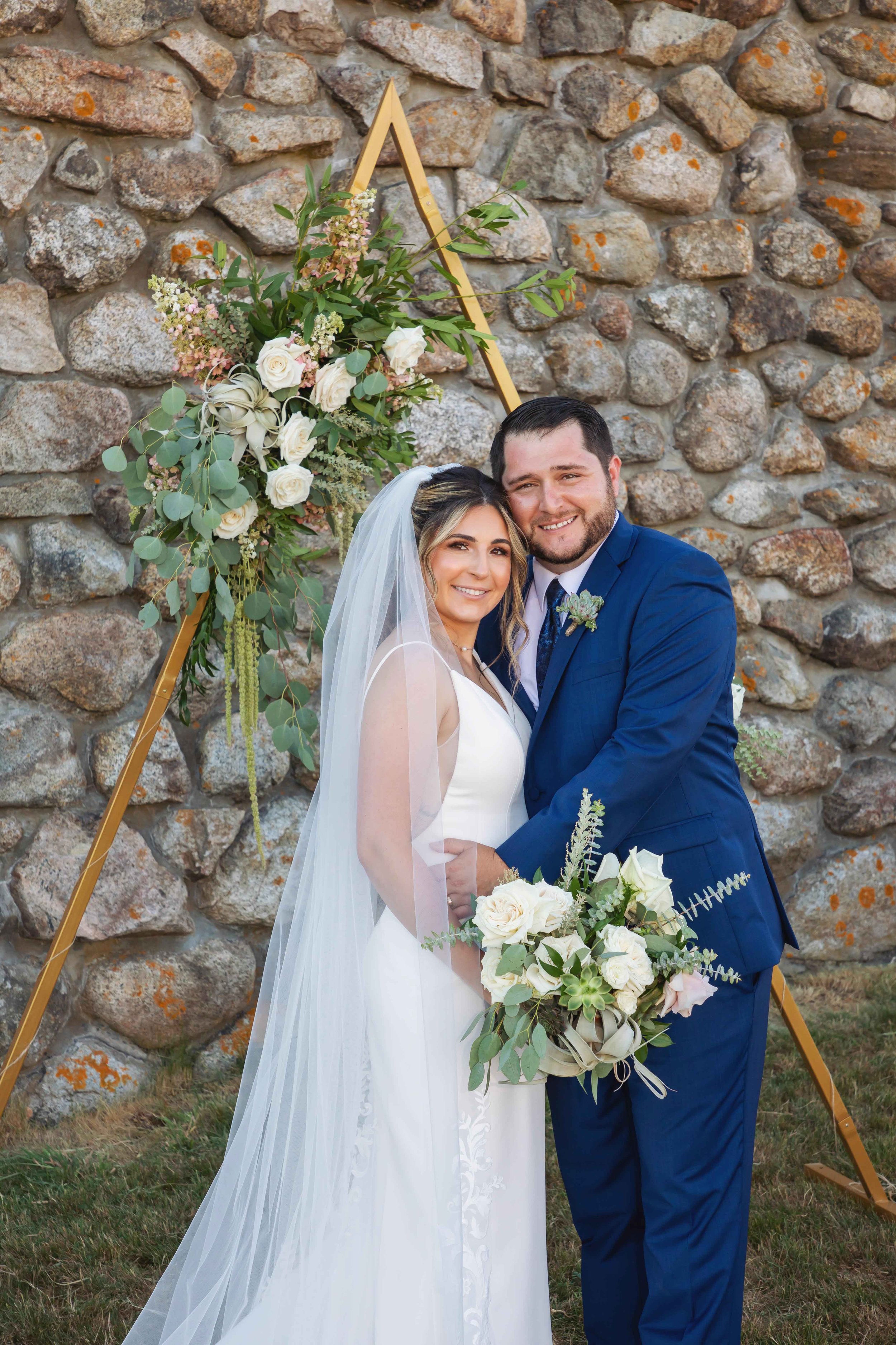 Bride in white wedding gown with veil and groom in blue suit, holding a bouquet of white roses and greenery, standing in front of a stone wall with a floral arrangement on a gold geometric arch.