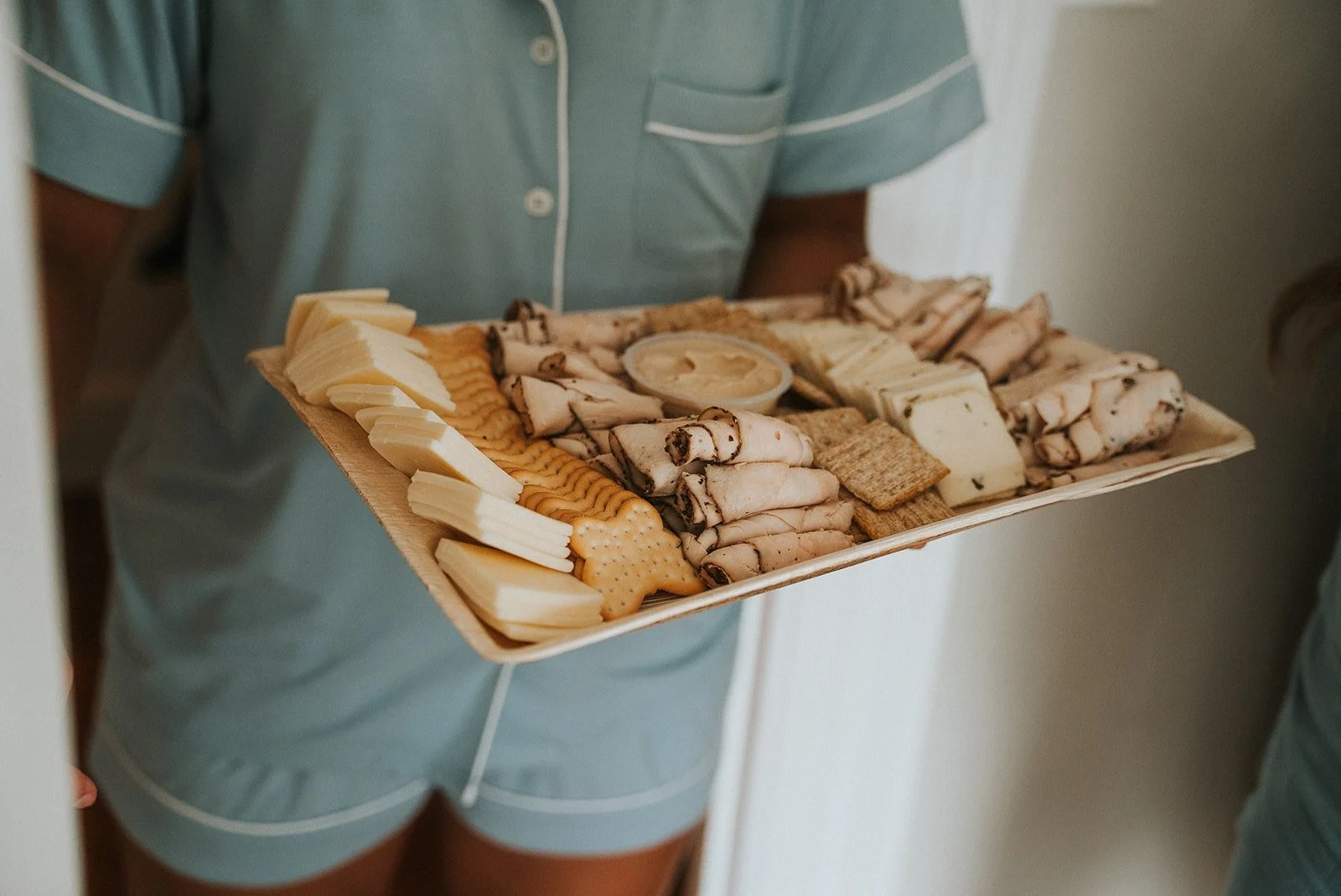 A person in blue pajamas holding a tray of assorted cheeses and crackers.