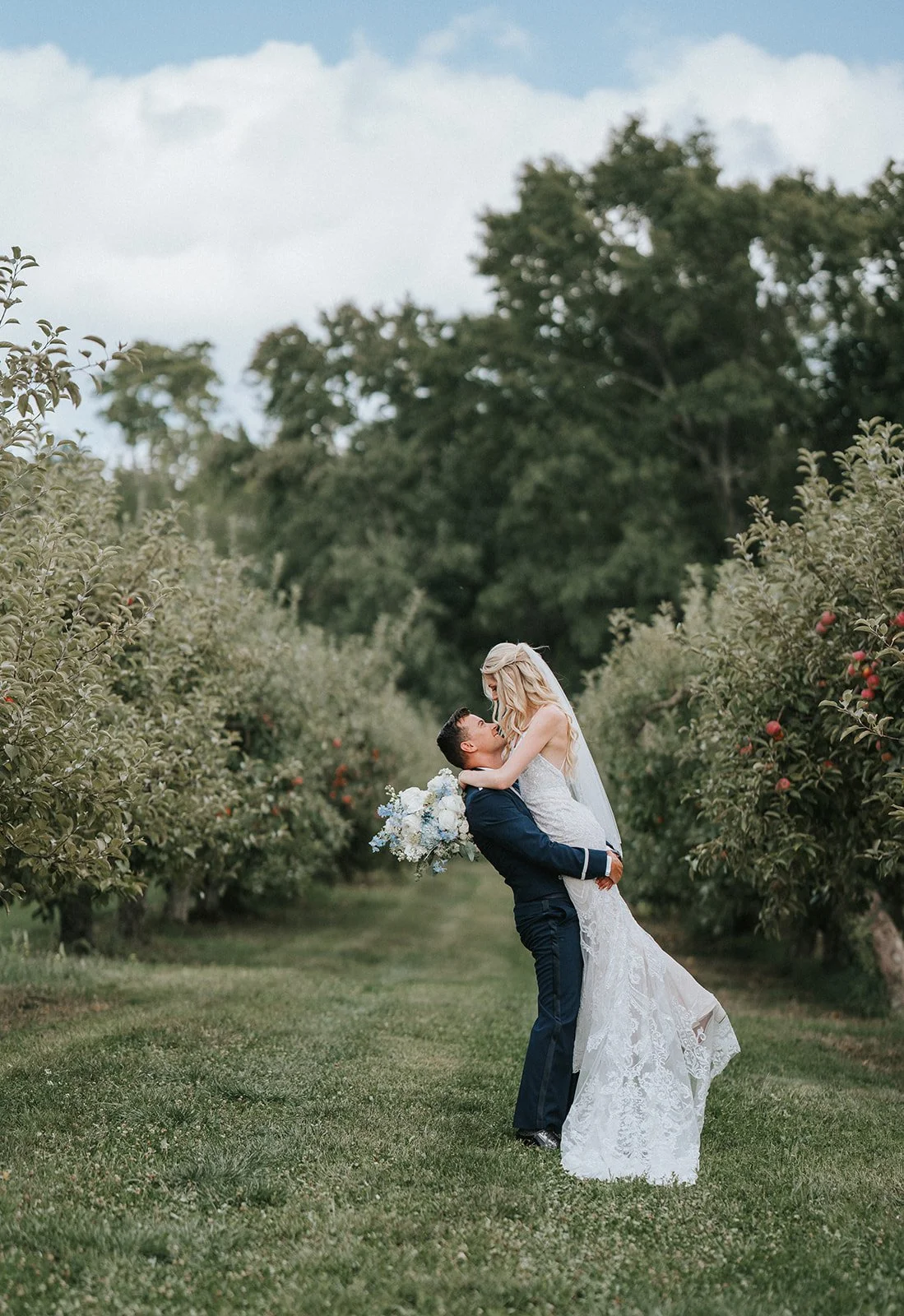 A newlywed couple outdoors in a garden, with the groom lifting the bride while she holds a bouquet of white flowers, surrounded by apple trees and lush greenery.
