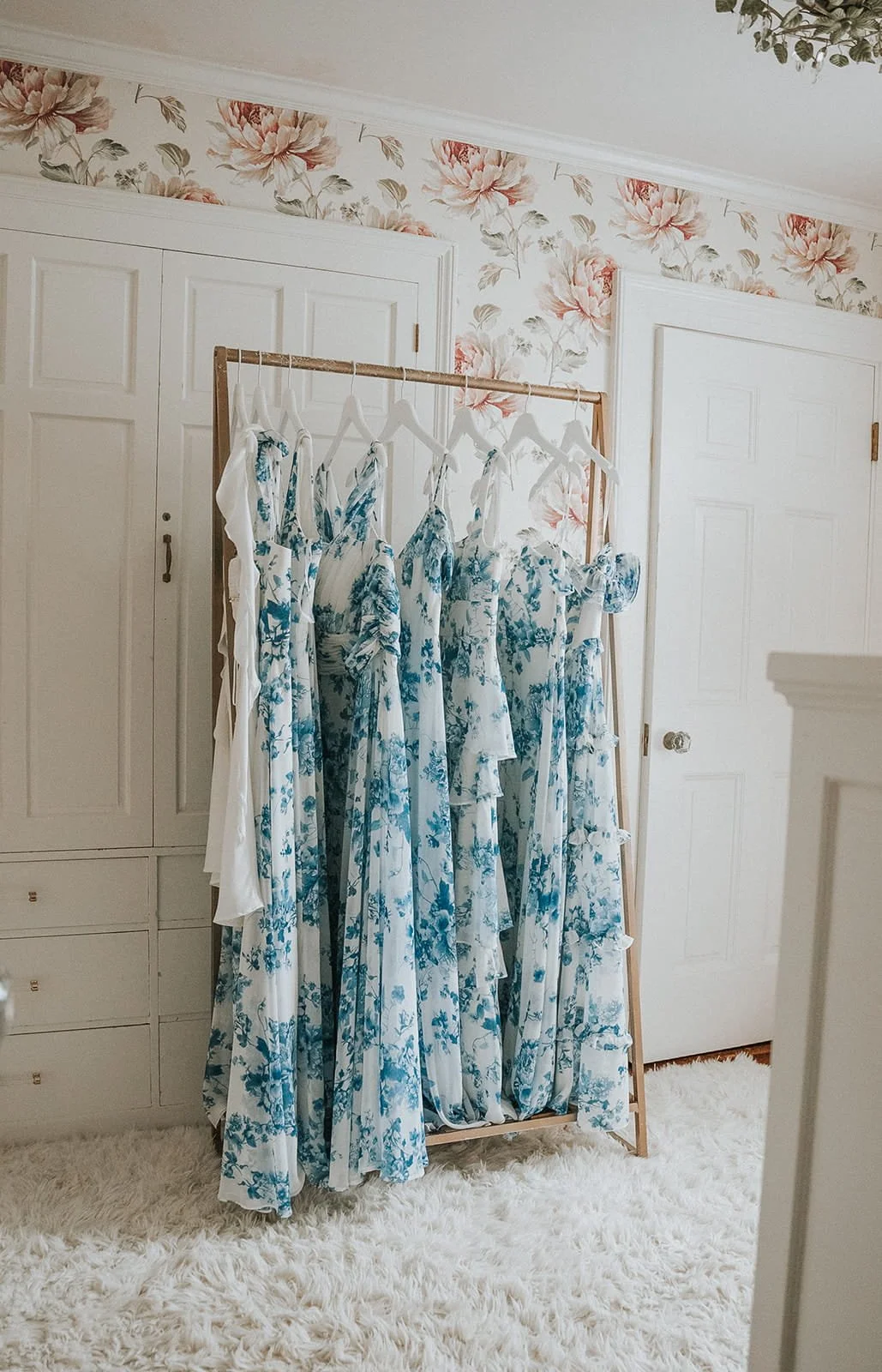 A clothing rack with blue floral dresses hanging in a room with floral wallpaper and a white, fluffy rug.