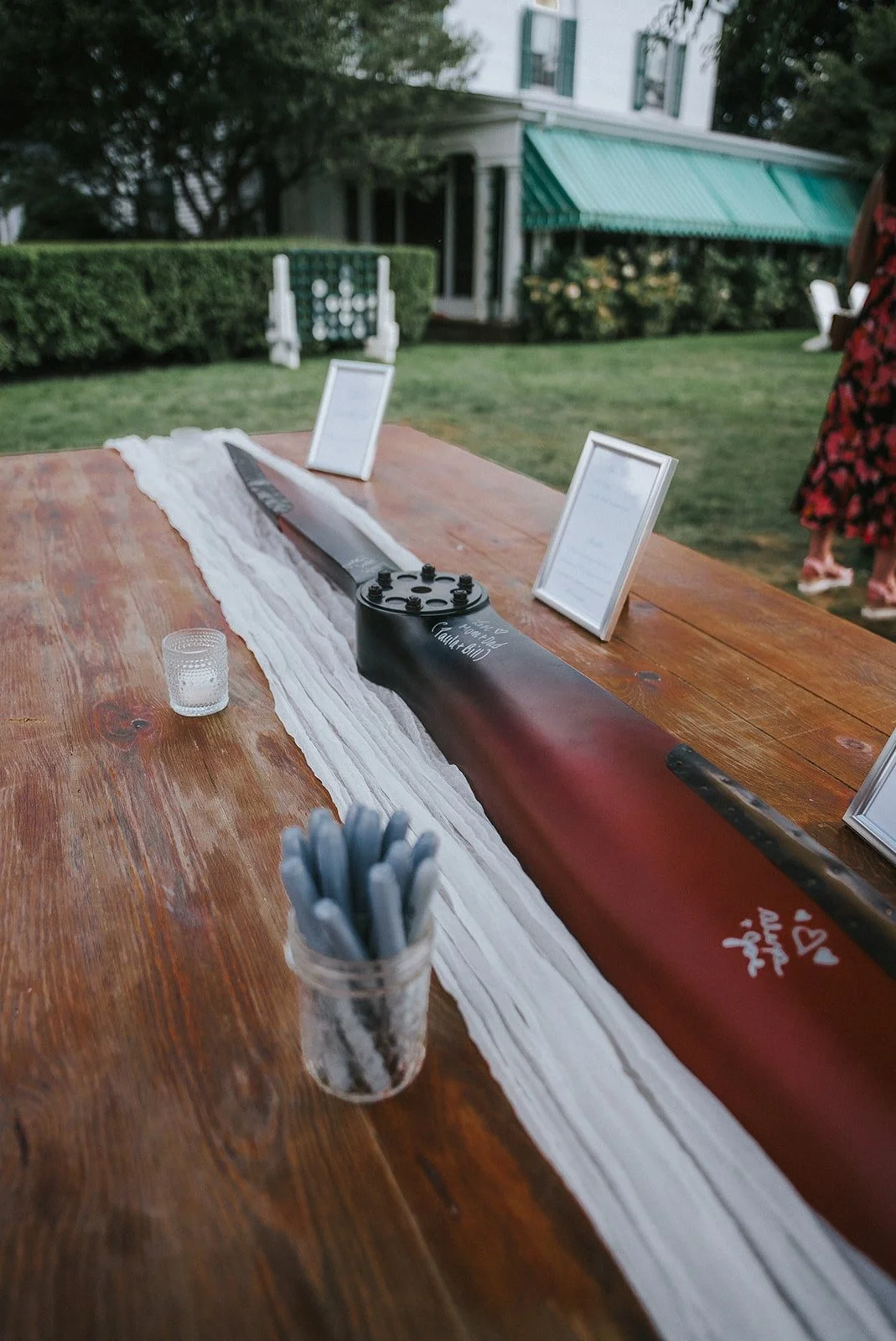 A long table with a white table runner, a toy lightsaber, framed signs, a small glass container with pens, and a small candle holder. In the background, there are people and a house with a green awning and a neatly trimmed lawn.