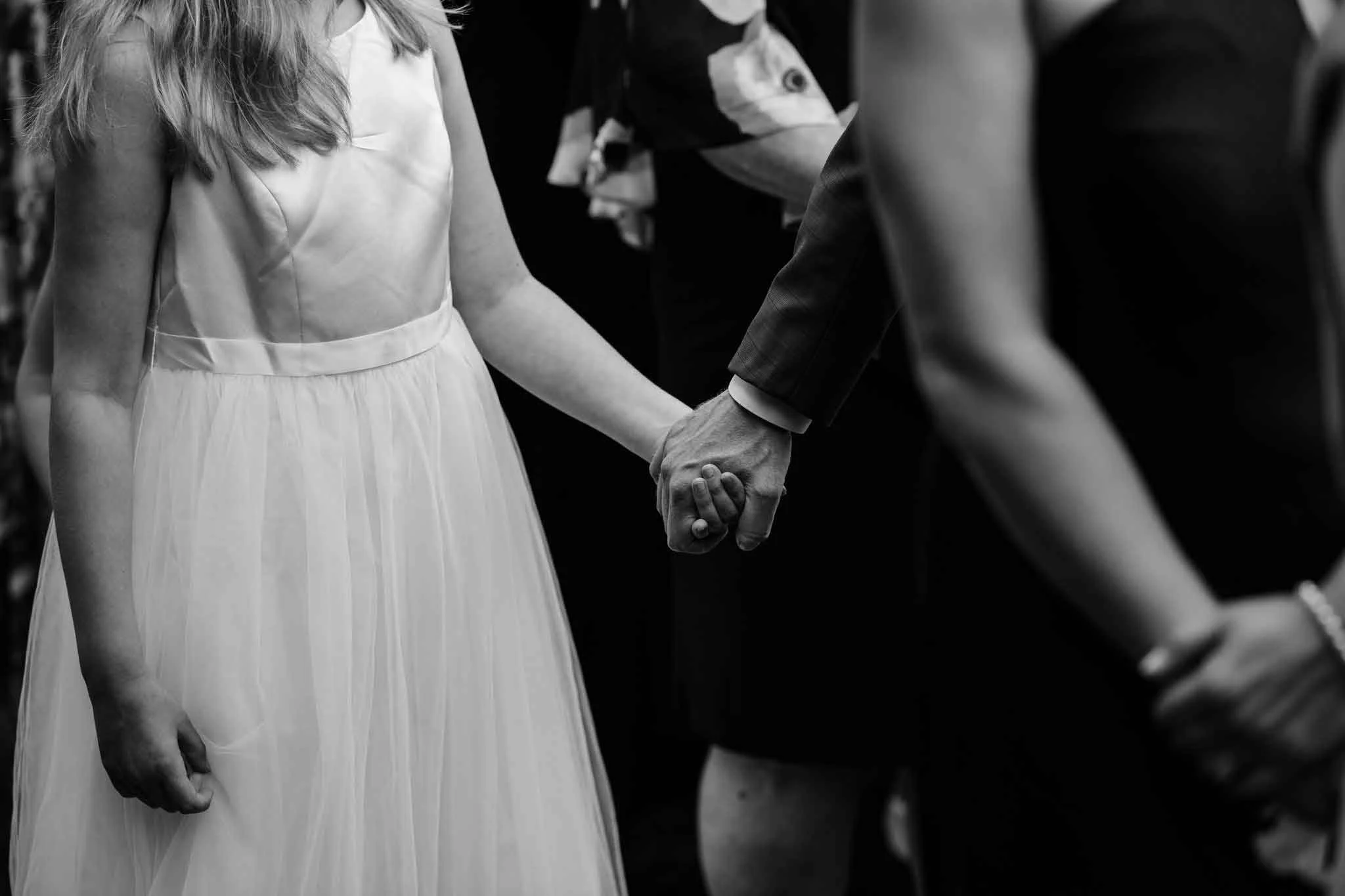People holding hands, including a woman in a white dress and a man in a suit, at a wedding ceremony.