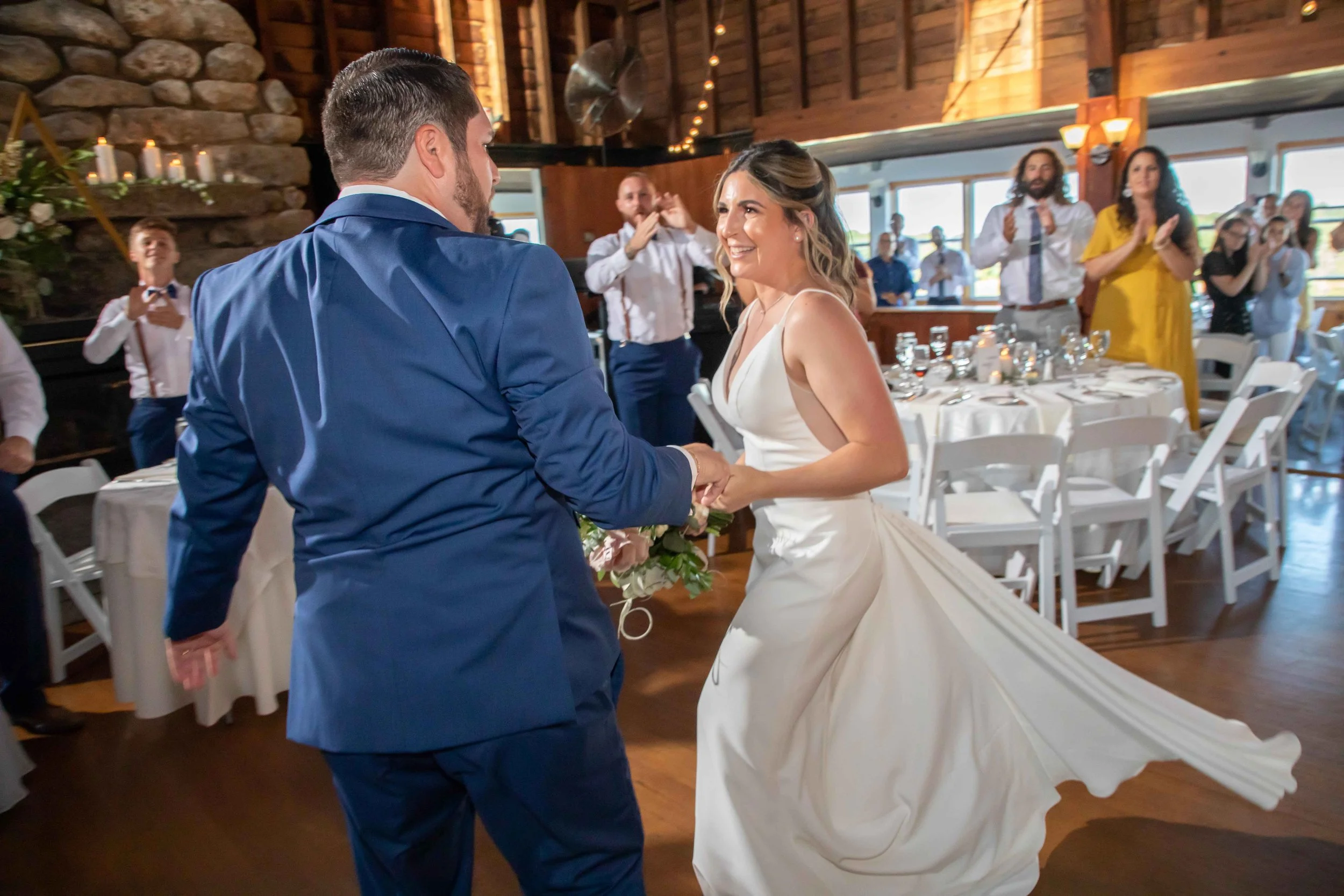 Bride and groom dancing at wedding reception in a rustic venue with friends clapping in the background.
