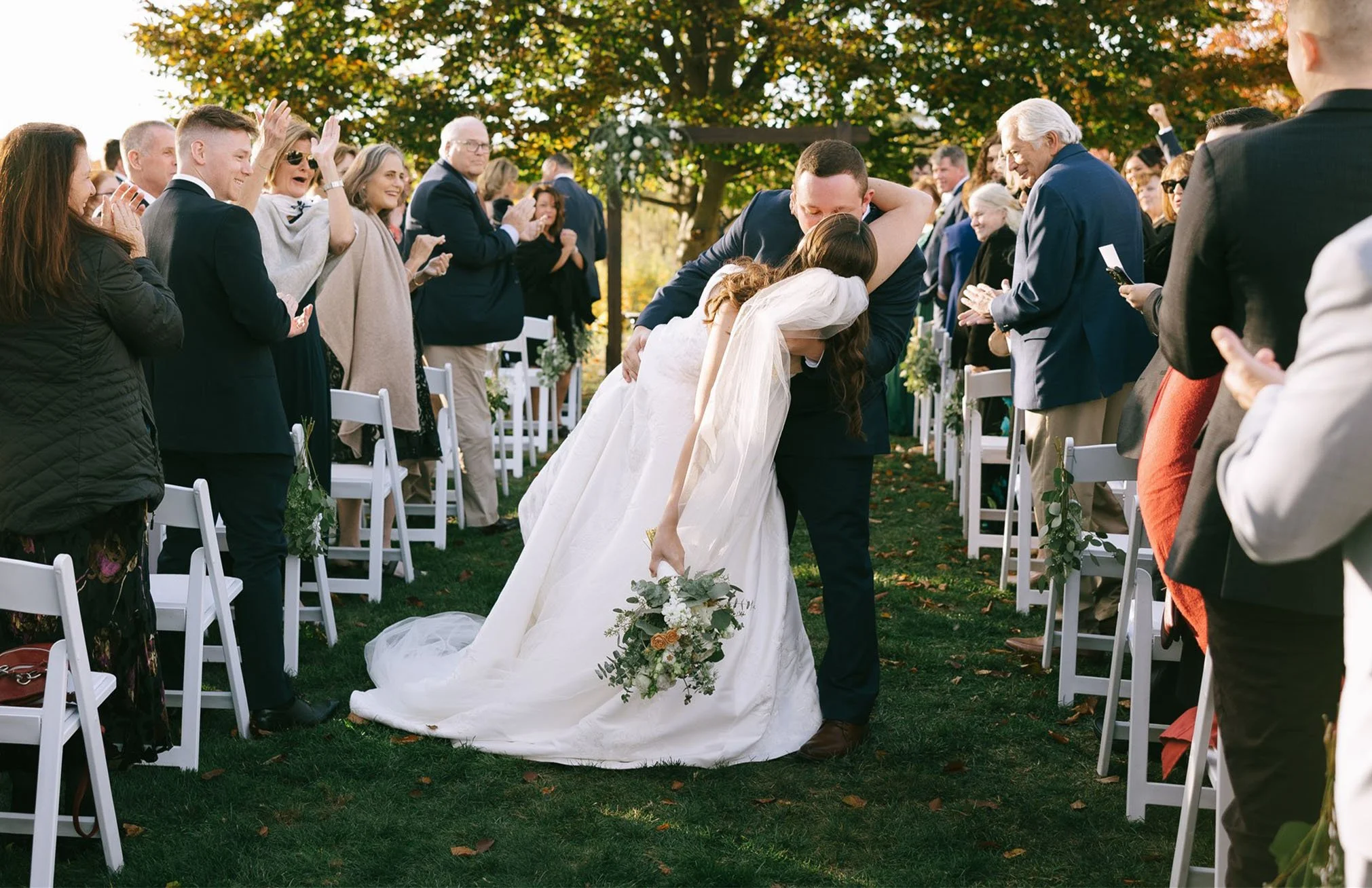 A bride and groom kiss while bowing at their wedding outdoors, surrounded by seated guests applauding under a tree with fall foliage.