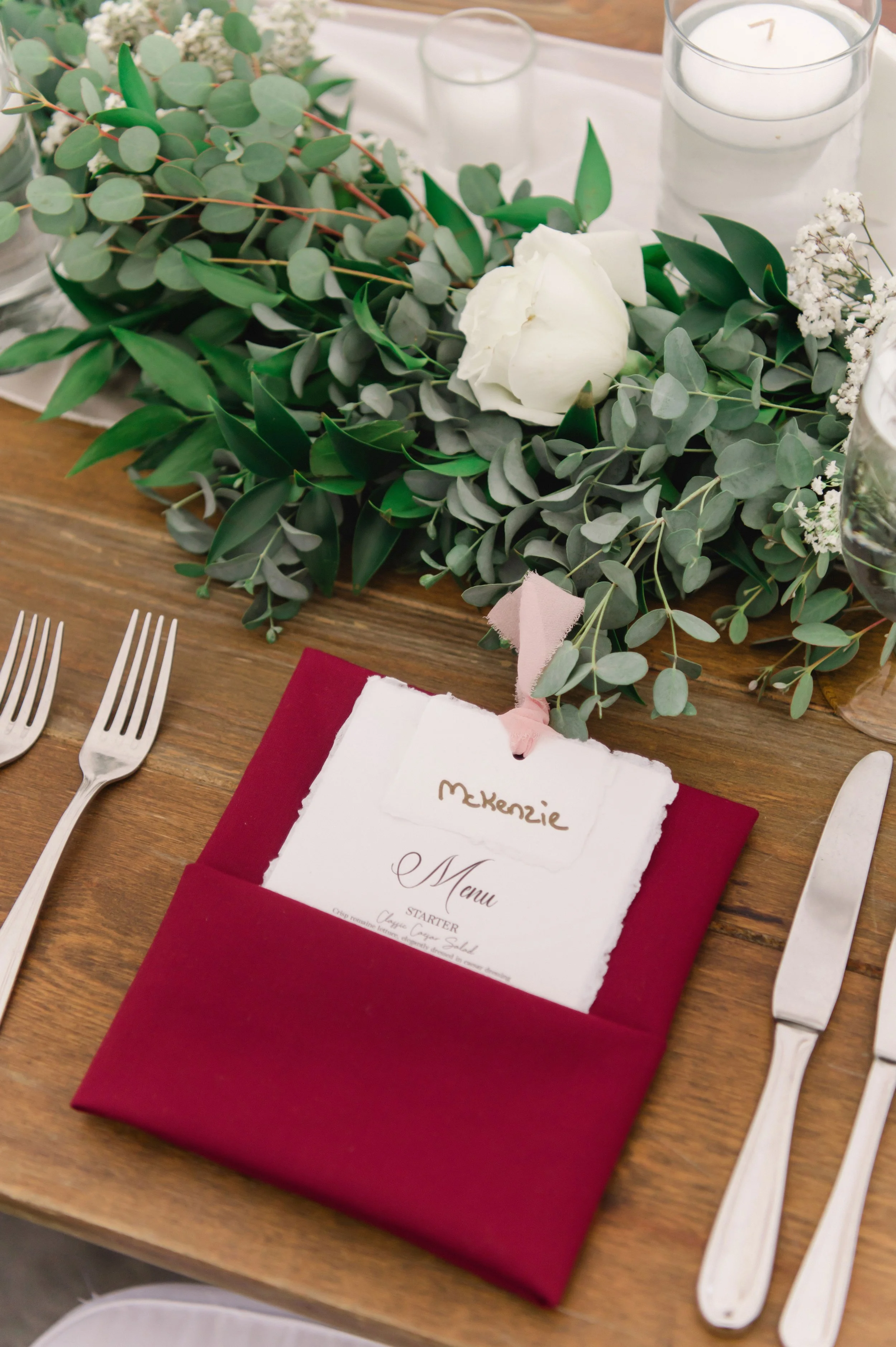 Table setting with a burgundy napkin holding a torn-edge menu with the name 'Mckenzie', surrounded by silverware and a floral centerpiece of eucalyptus leaves, white roses, baby's breath, and candles.
