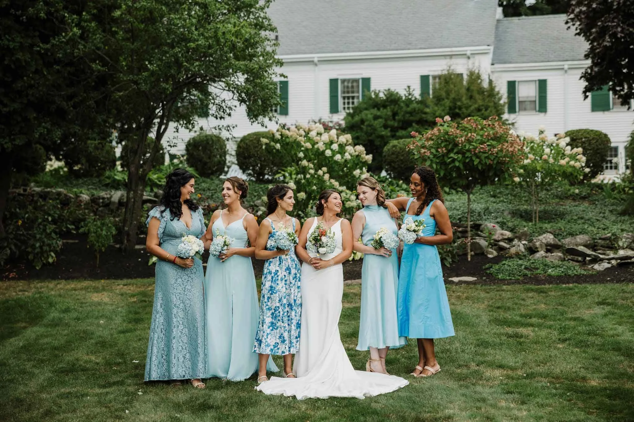 Group of six women dressed in pastel and blue dresses, holding bouquets of flowers, standing on a grassy lawn with trees and a white house in the background, smiling and engaging with each other