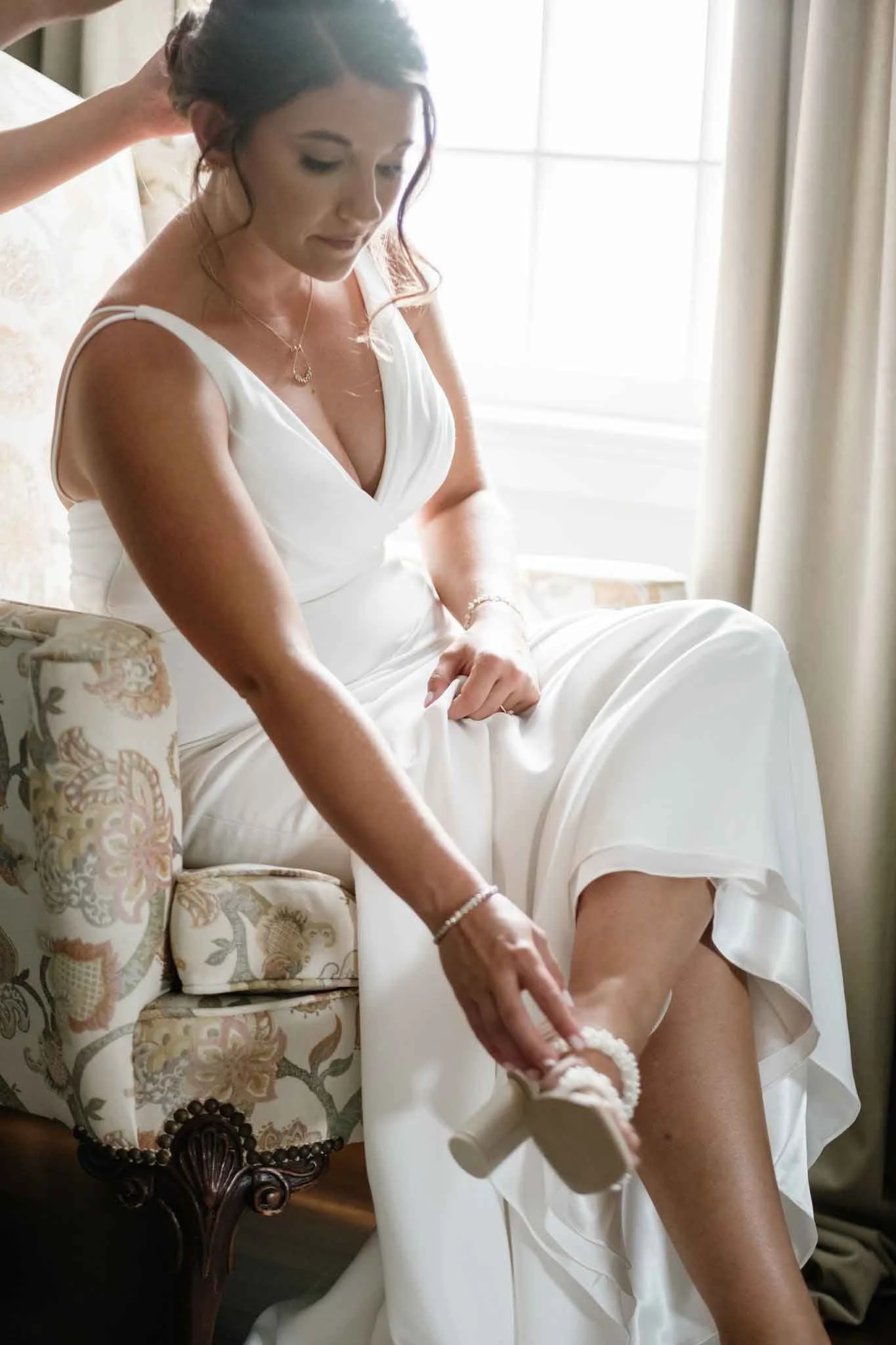 A woman in a white dress sitting on a floral upholstered chair, adjusting her high-heeled sandal, with a window and curtains in the background.