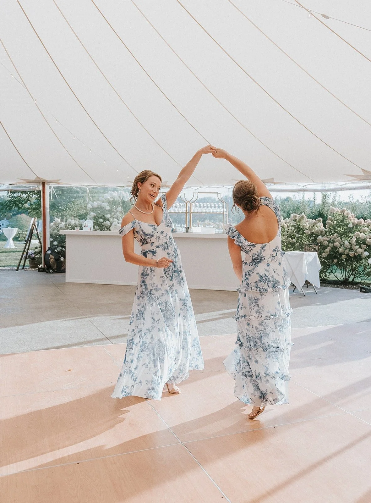 Two women in matching blue and white floral dresses dancing in a tent during a celebration or wedding reception.