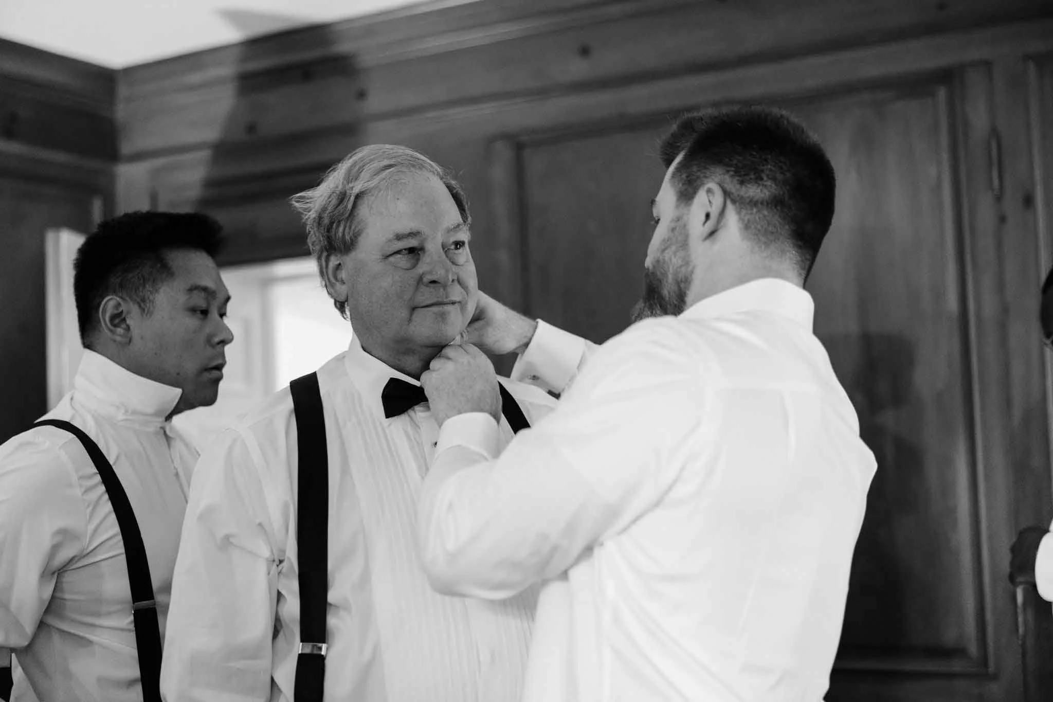 Men dressing formally, with one adjusting another's bow tie, in a wooden-paneled room.