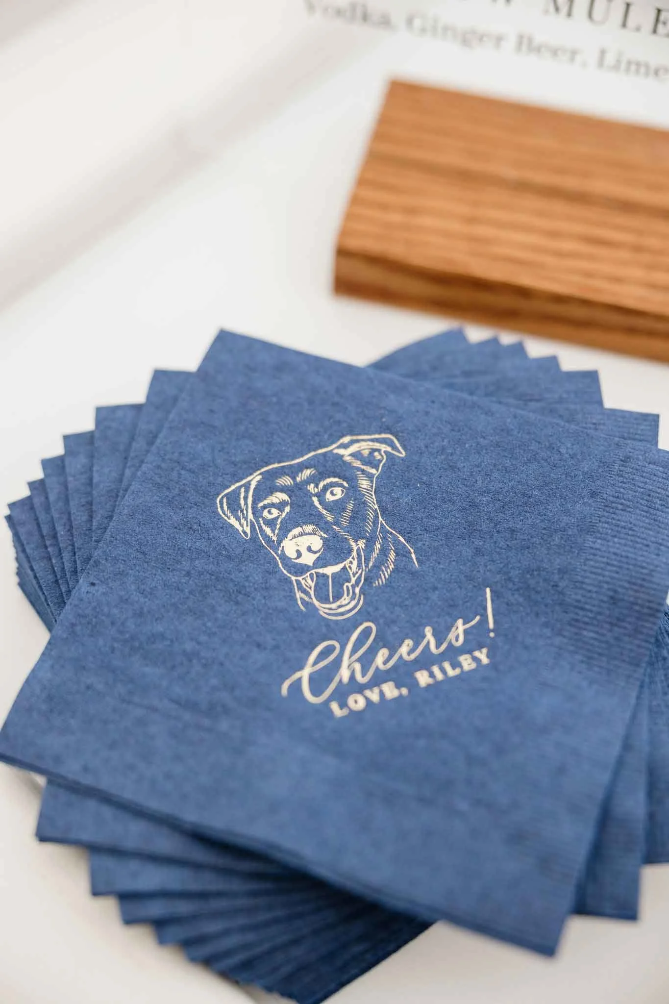 A stack of blue paper napkins with a white line drawing of a dog's face, the word "Cheers!" and the phrase "Love, Rilby" written on them.