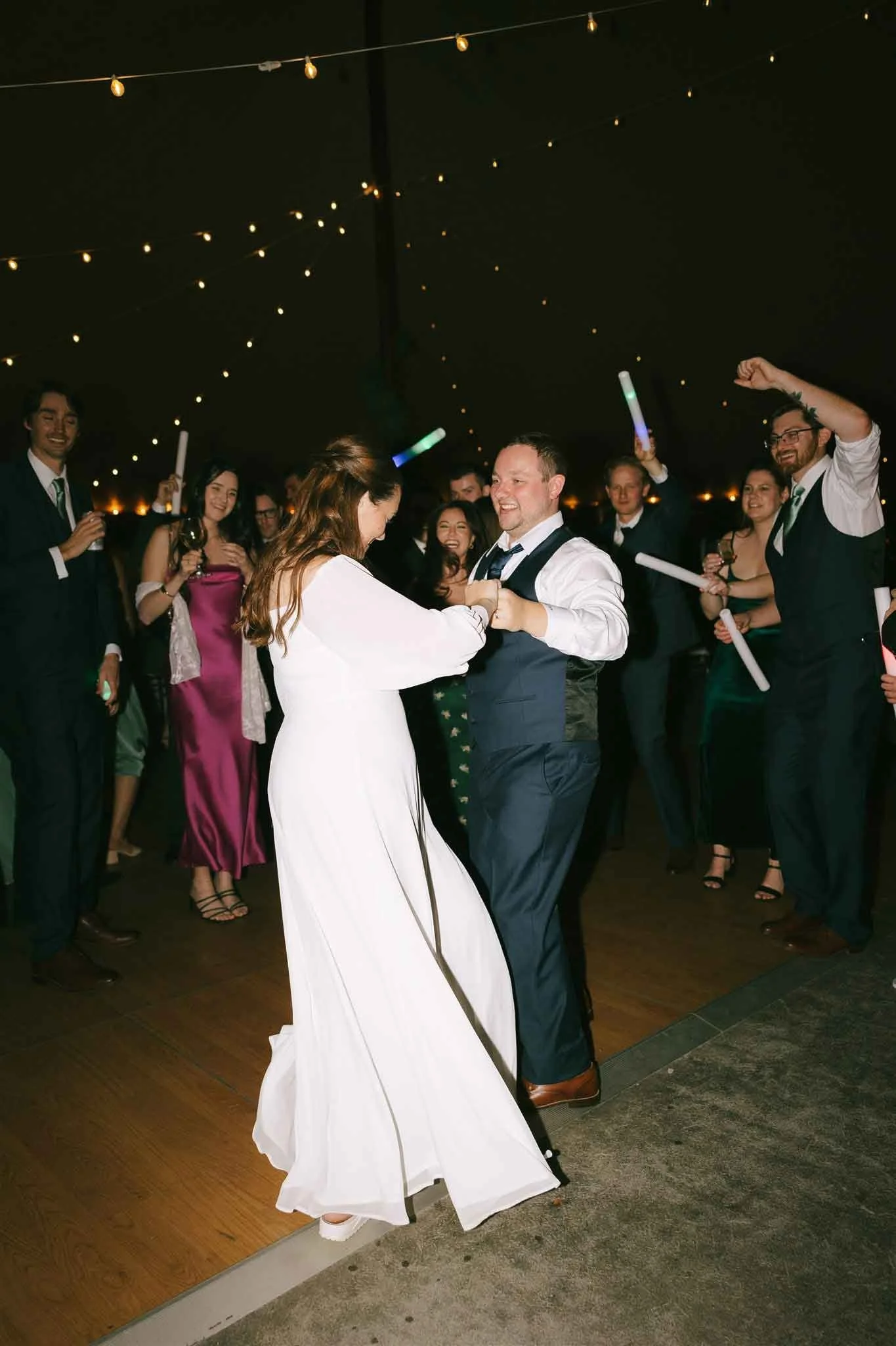 A bride and groom are dancing in the center of a wedding reception while guests surround them, celebrating with glow sticks and drinks under string lights.