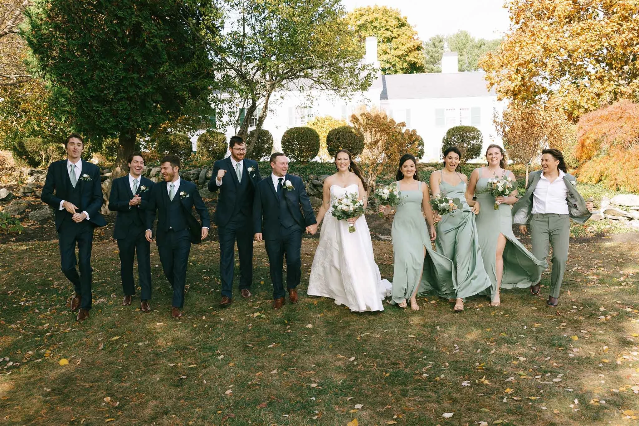 A wedding party walking outdoors on a grassy area with trees and a white house in the background. The group includes five men in suits, a bride in a white wedding gown, and five women in matching light green dresses, all holding bouquets of flowers.