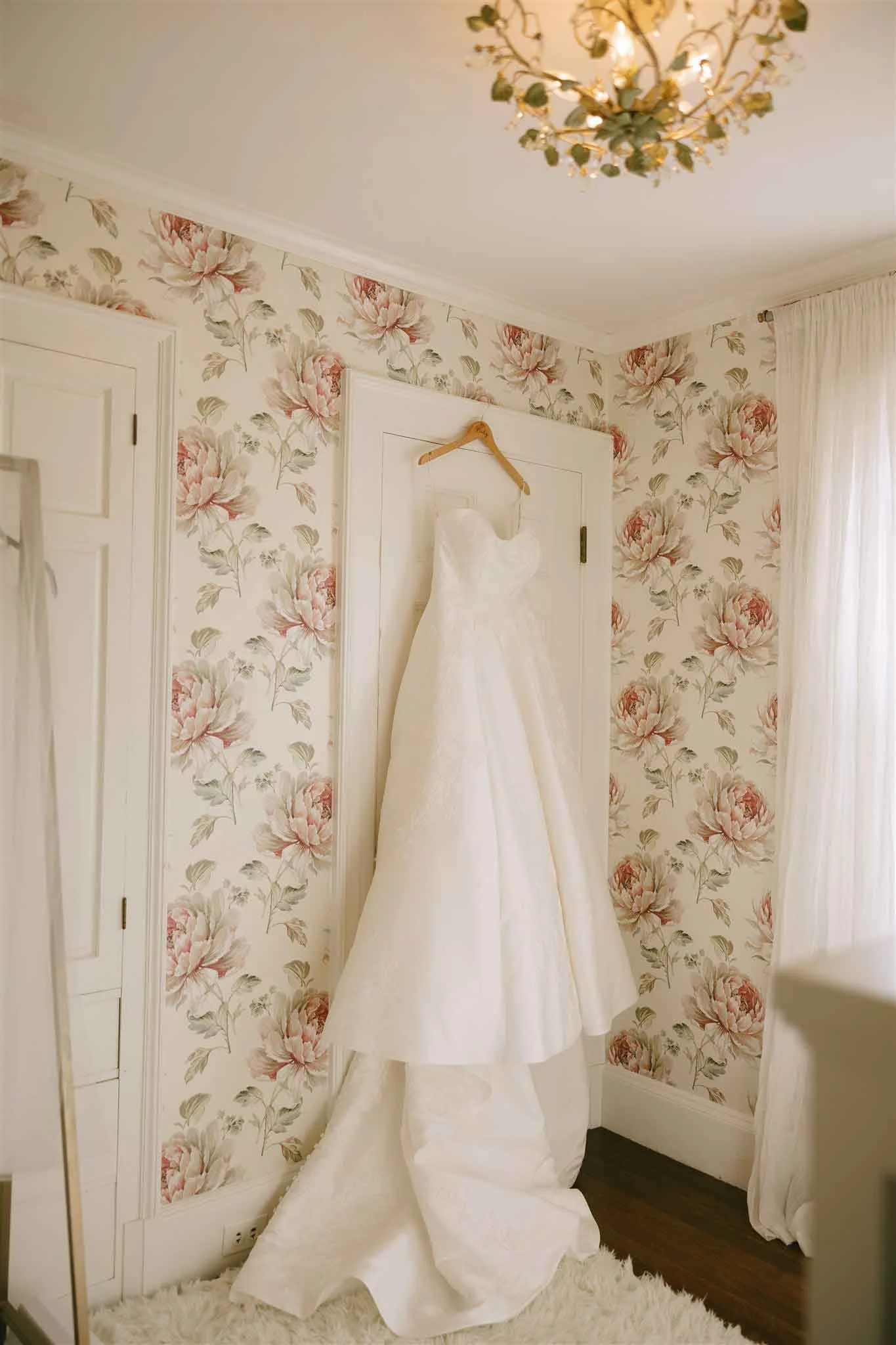 A wedding dress hanging on a hanger on a door in a room with floral wallpaper and a chandelier.