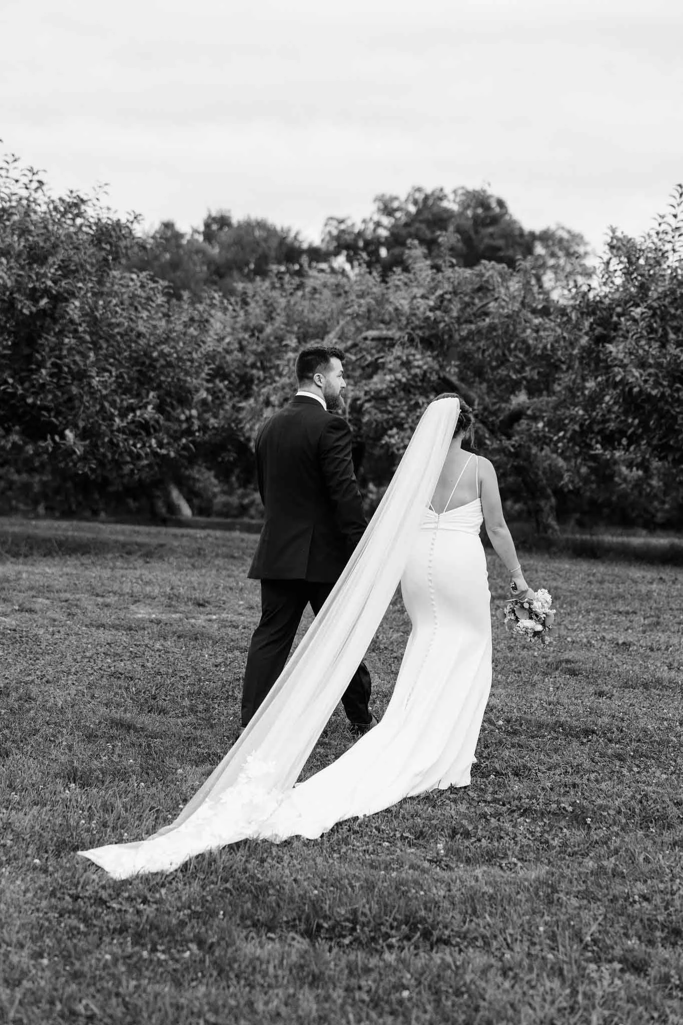 Black and white photo of a bride and groom walking outdoors on grass, with bushes and trees in the background. The bride is wearing a long white gown with a train and a veil, holding a bouquet. The groom is in a dark suit.