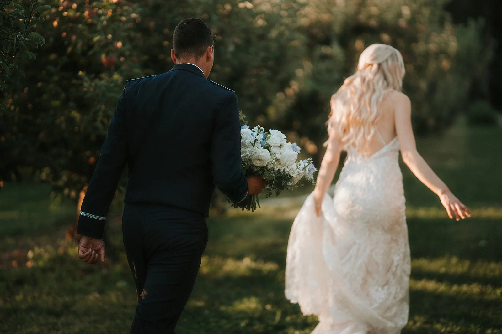 A groom holding a bouquet of white flowers walking behind a bride in a white lace wedding dress with long curls in an outdoor setting with greenery.