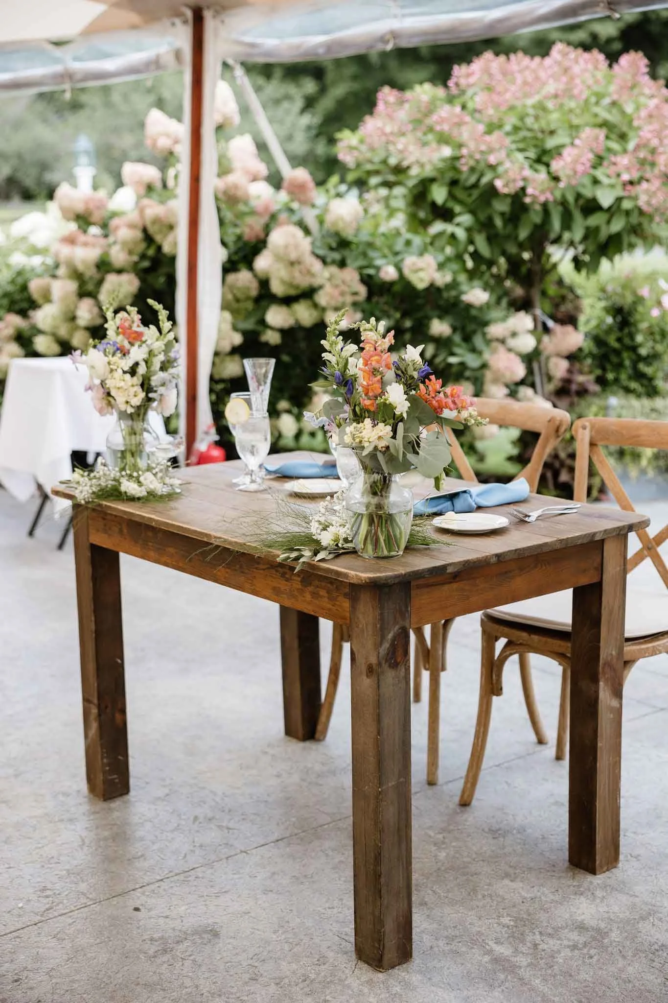 A rustic wooden dining table set outdoors with floral centerpieces, surrounded by wooden chairs, with pink and white hydrangeas blooming in the background.