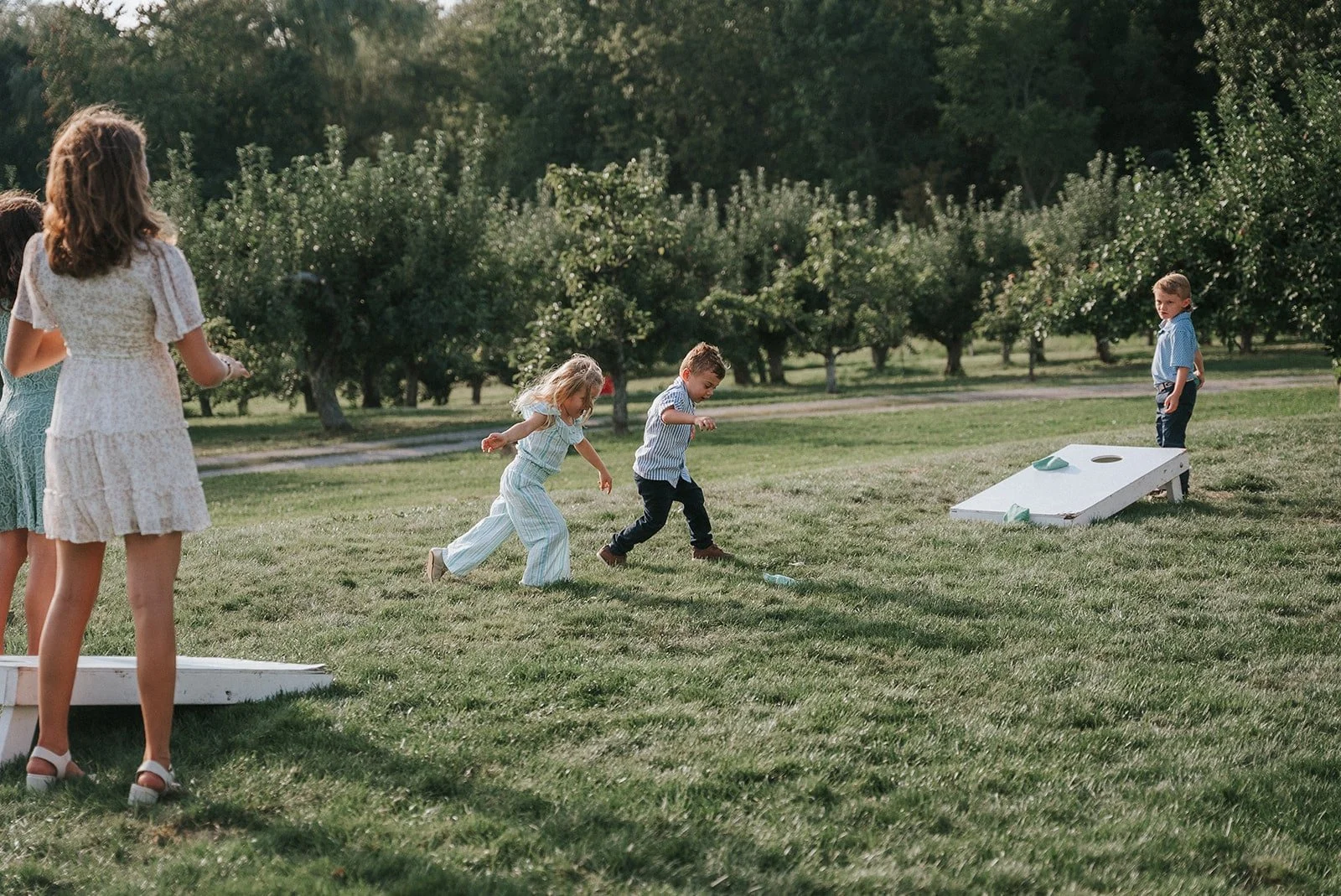 Children playing a bean bag toss game outdoors on a grassy field with trees in the background.