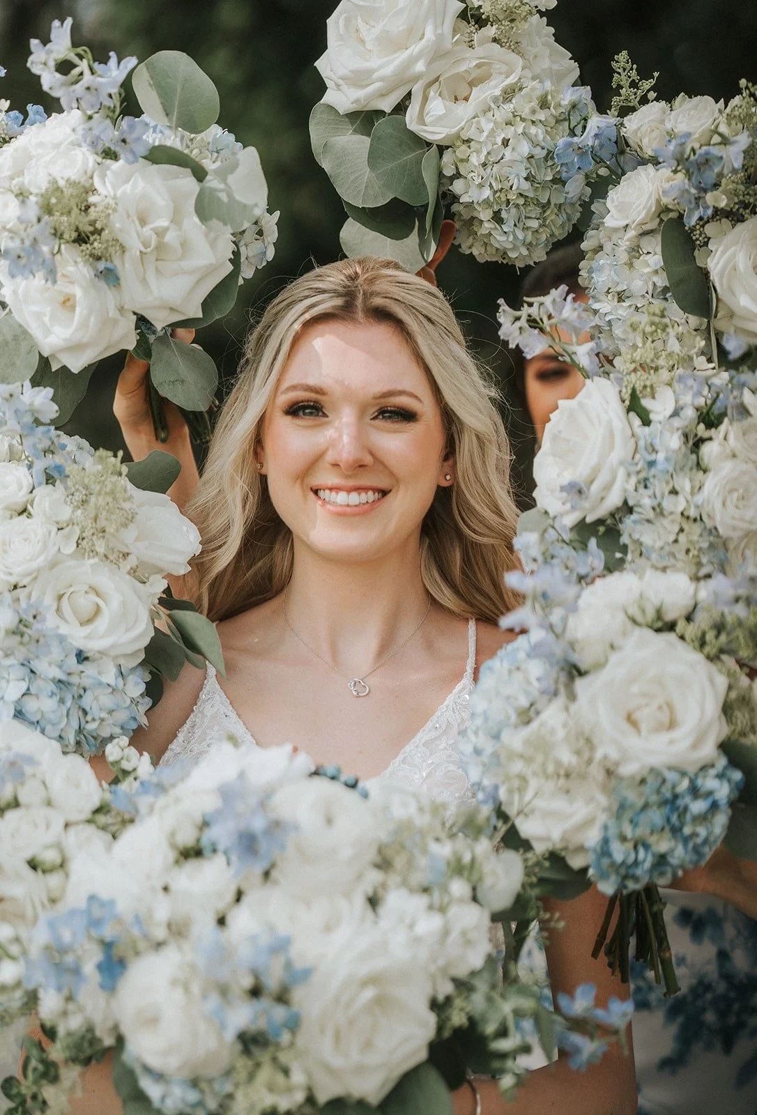 A smiling woman with long blonde hair in a white dress surrounded by a floral arch of white roses, blue hydrangeas, and eucalyptus leaves.
