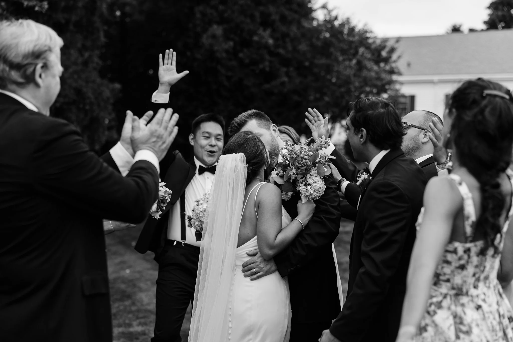 Black-and-white photo of a newlywed couple kissing, surrounded by friends and family celebrating outdoors with raised hands and holding bouquets.