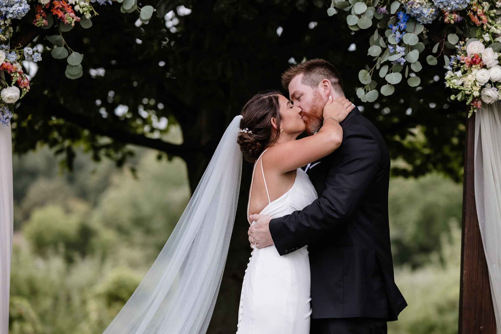 A bride and groom kiss during their outdoor wedding ceremony under a floral arch.