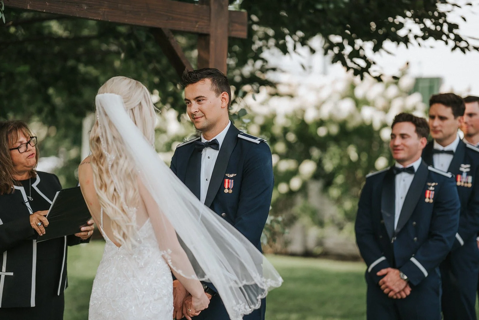A wedding ceremony outdoors with a bride and groom exchanging vows, surrounded by groomsmen in military uniforms and an officiant.