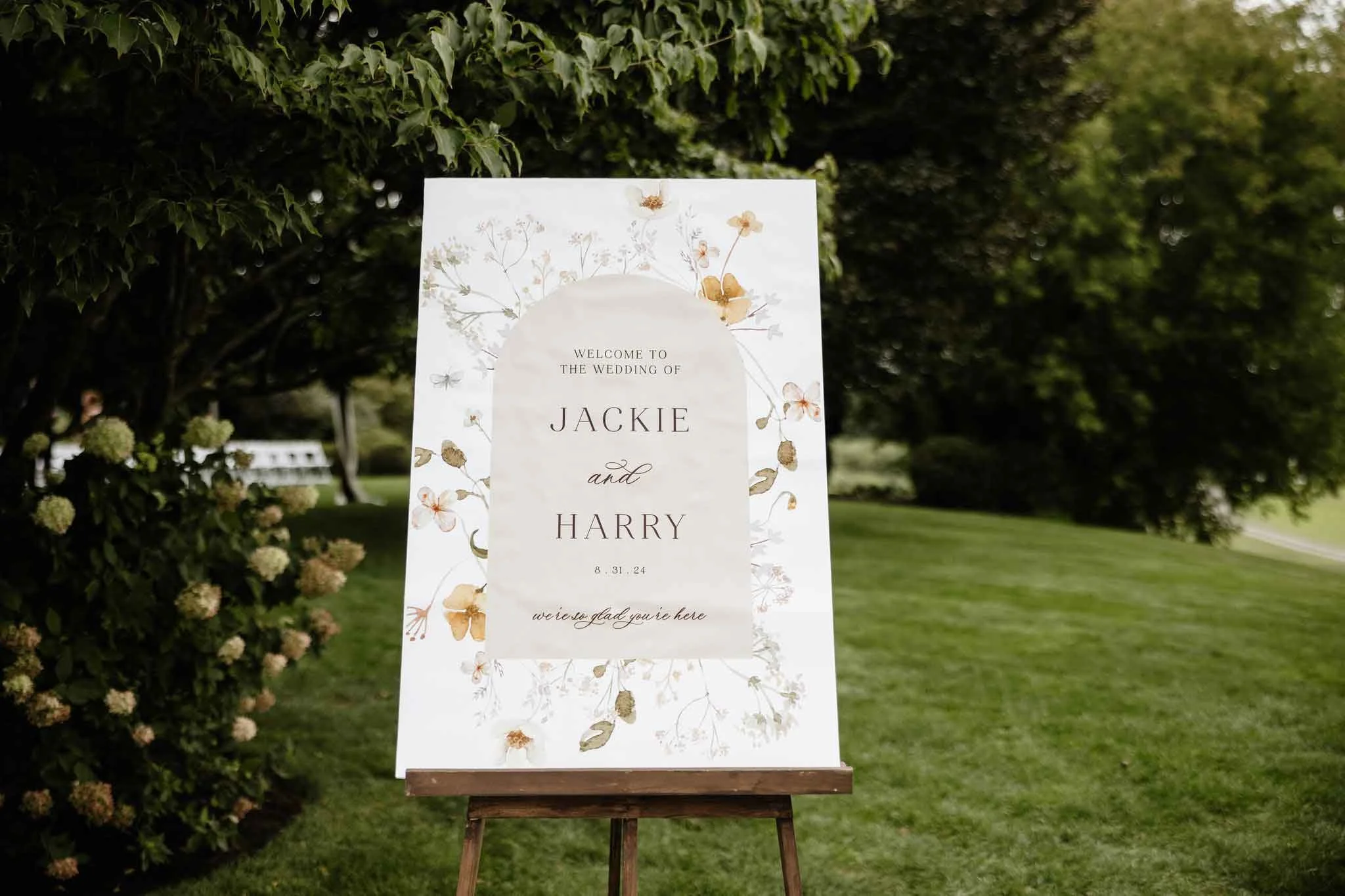 Wedding welcome sign on an easel outdoors surrounded by greenery, with floral designs and text welcoming guests to the wedding of Jackie and Harry, dated August 31, 2024.