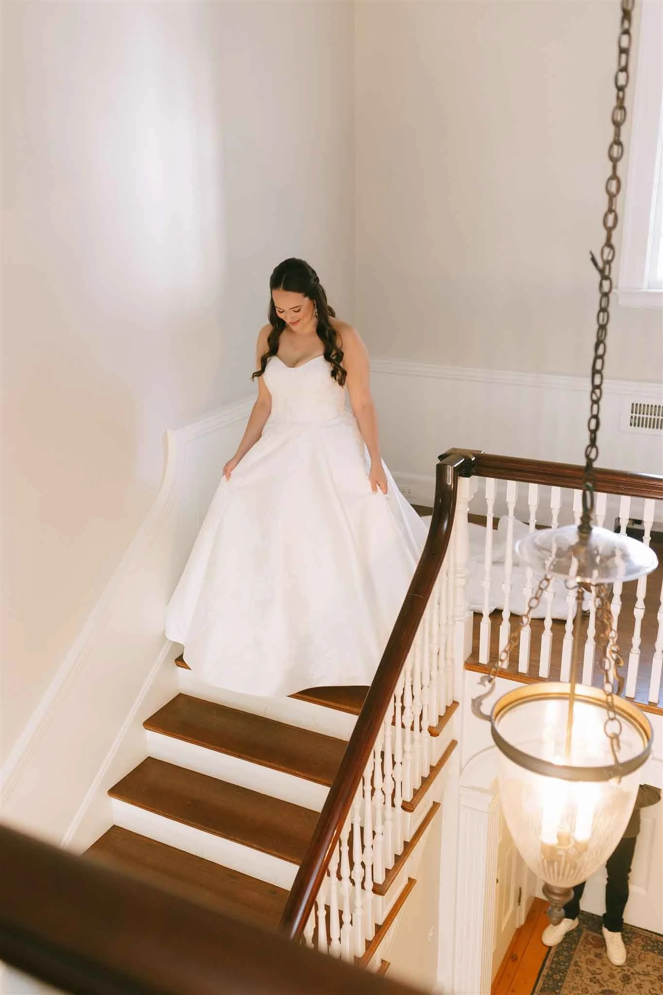 A bride in a white wedding dress standing at the top of a staircase, looking down, with a man partially visible at the bottom of the stairs.