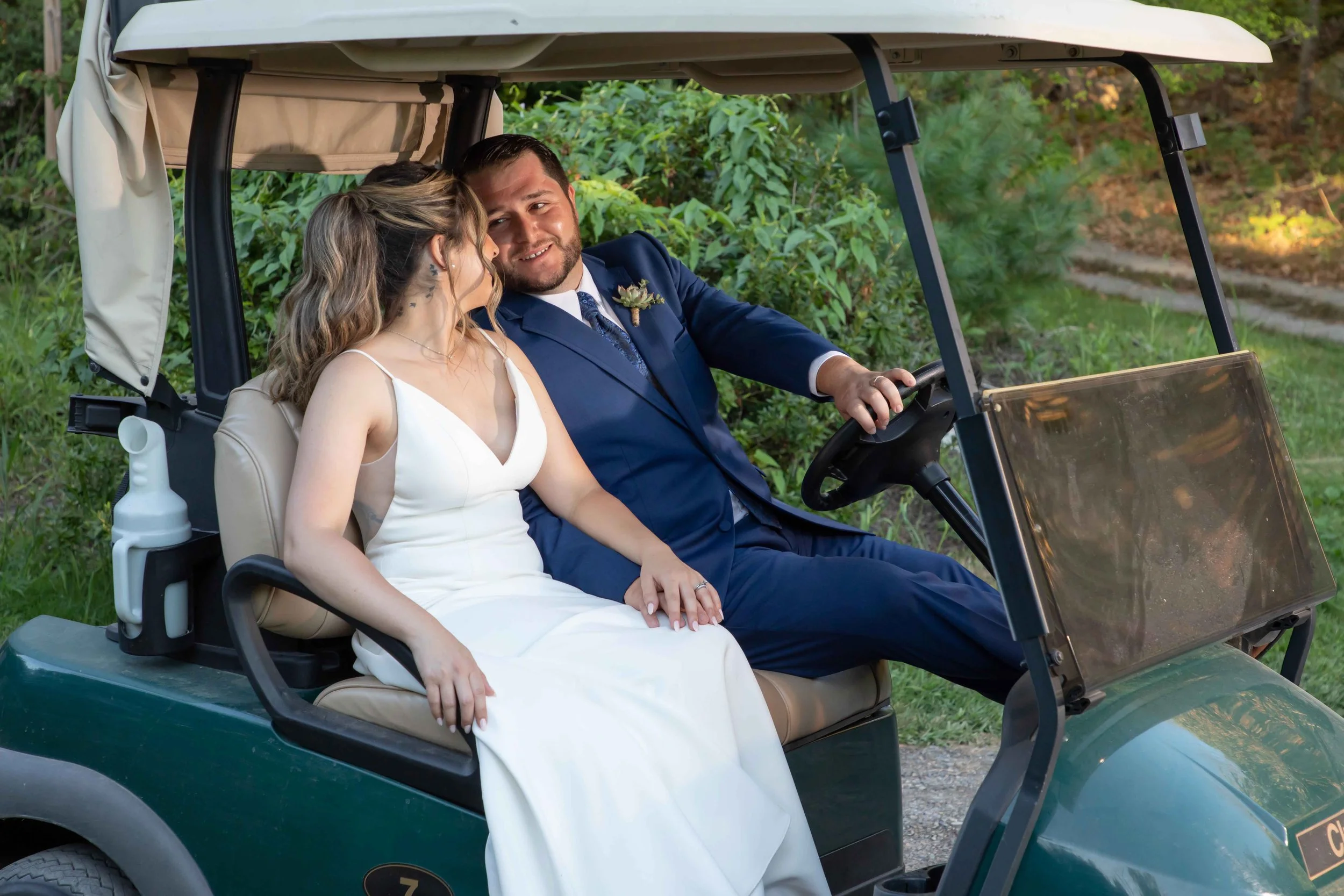 A bride and groom sit together in a golf cart on a grassy area surrounded by greenery, smiling and sharing a tender moment during their outdoor wedding.
