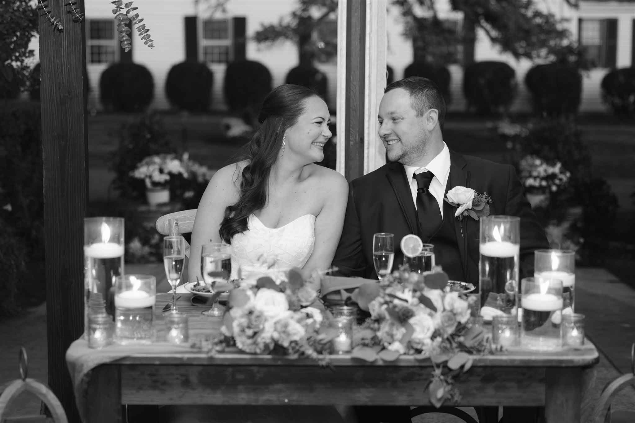 Black and white photo of a bride and groom at their wedding reception, sitting at a decorated table with candles and flowers, smiling at each other.