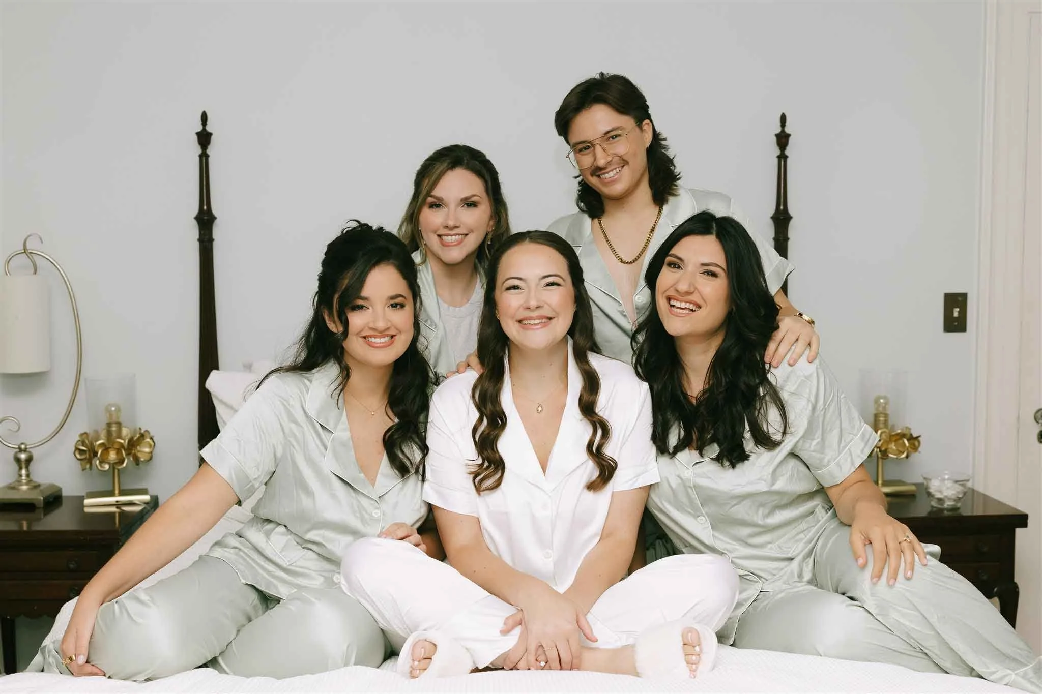 Six young women smiling in pajamas sitting on a bed in a bedroom.