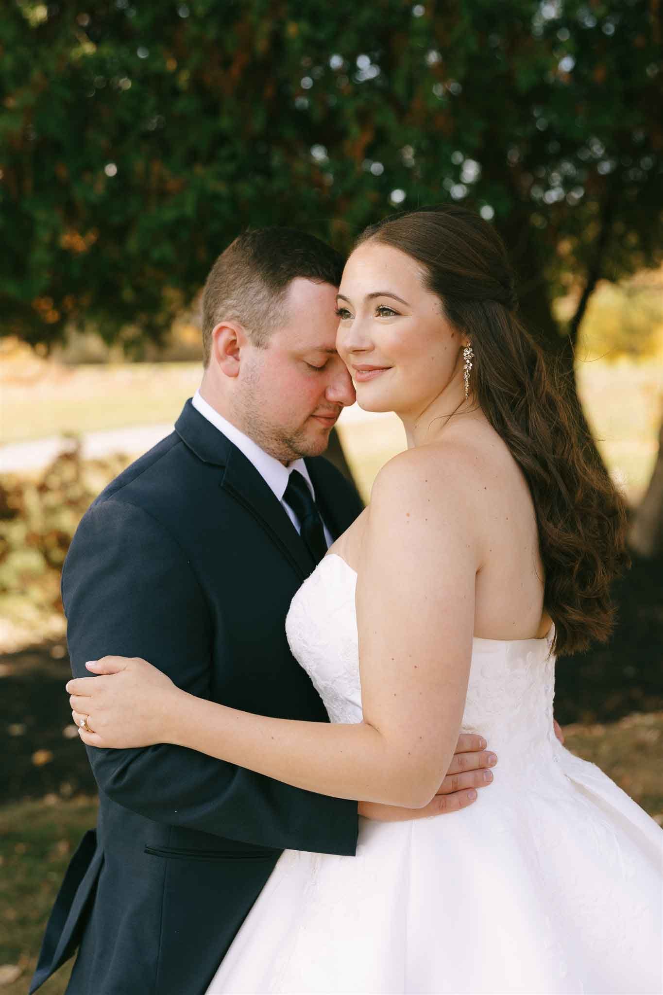 A bride and groom embrace outdoors during their wedding, with trees in the background. The bride is smiling, wearing a strapless white wedding gown with earrings, and has long, wavy brown hair. The groom, dressed in a dark suit and tie, has his eyes 