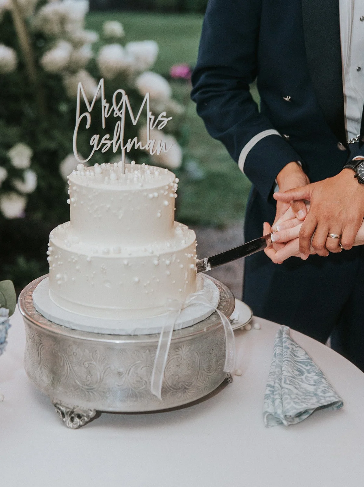 A newlywed couple cutting a two-tiered white wedding cake decorated with small white pearls. The cake has a topper that says 'Mr & Mrs Cashman'. The couple is holding a cake knife together, and the wedding is outdoors with white flowers in the background.