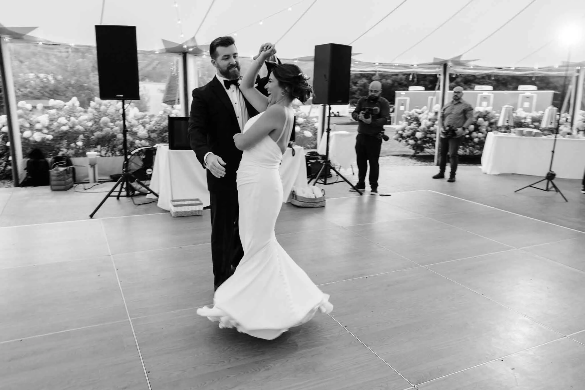 A bride and groom are dancing together at their wedding reception inside a decorated tent, with photographers and guests in the background.