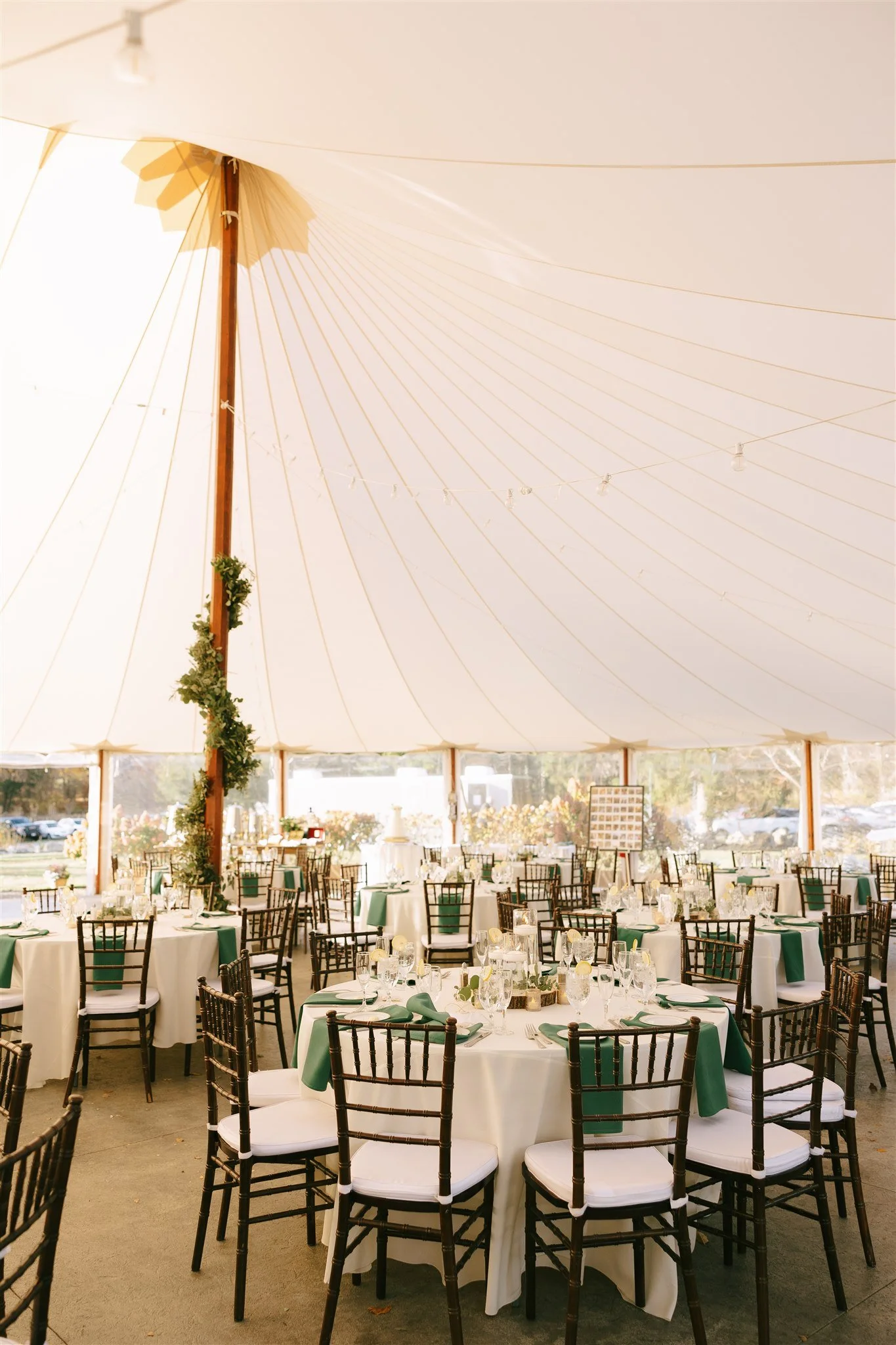 Wedding reception under a large white tent with round tables, green napkins, and wooden chairs set for a formal event.