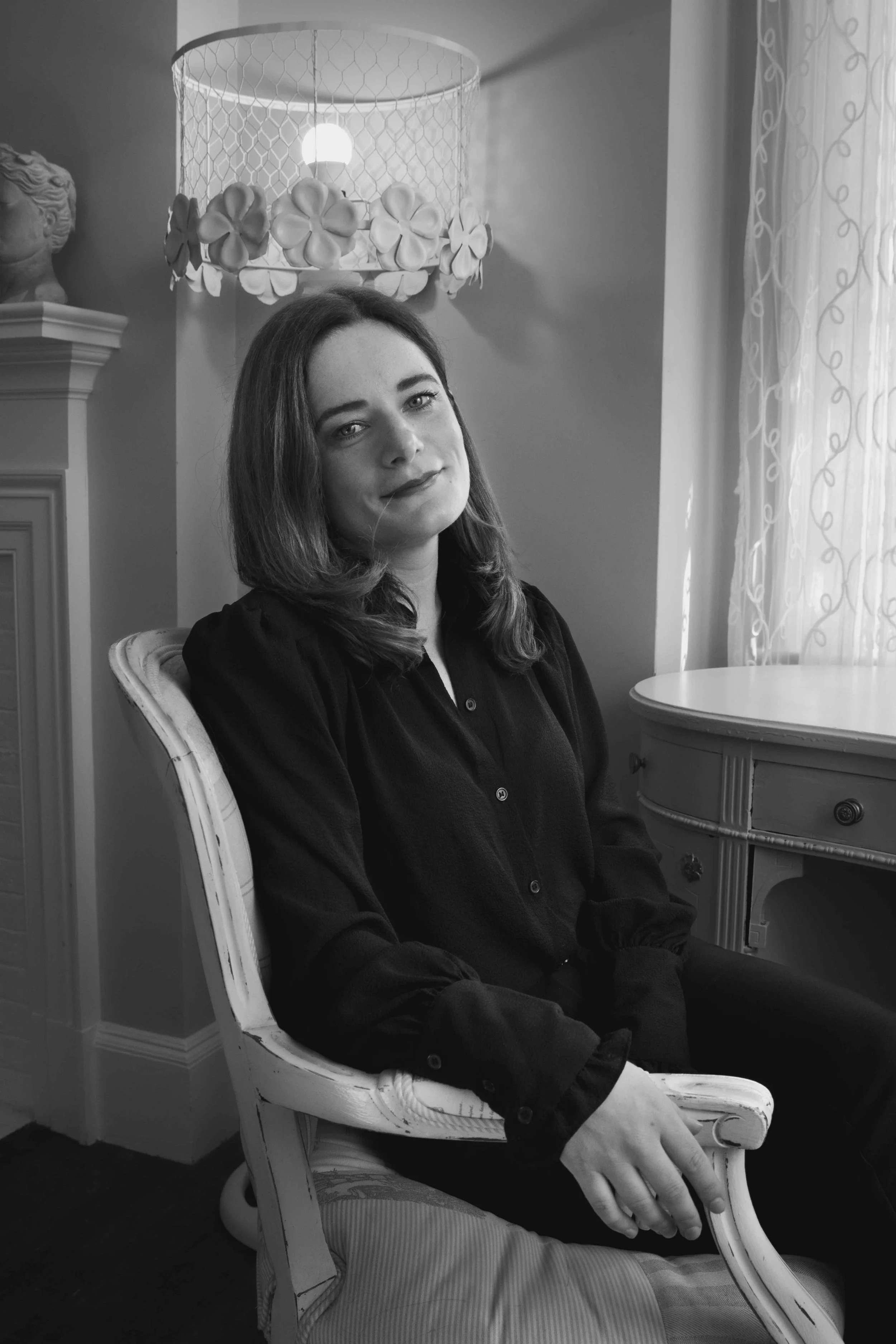 A black and white photo of a young woman with shoulder-length hair sitting on a vintage chair in a well-lit room with curtains and decorative lamps.